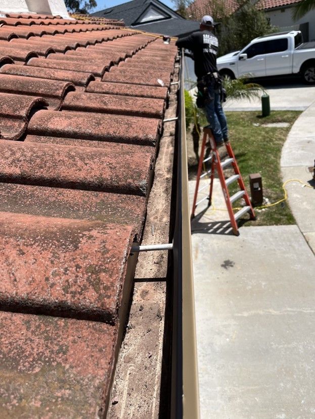 A person on a ladder cleans a roof gutter, with clay tiles, in an outdoor residential setting.