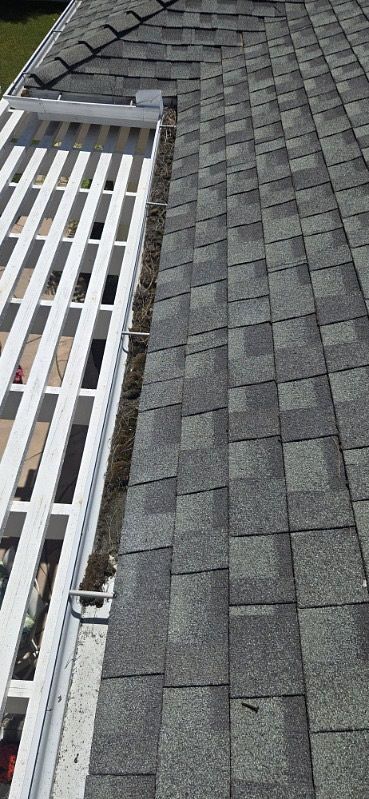A section of a roof with gray shingles and a white gutter guard. Debris is visible in the gutter.
