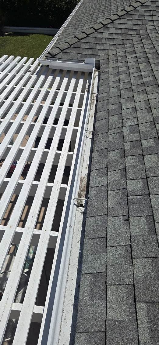 Overhead view of a roof with asphalt shingles and a white, slotted gutter guard system. The gutter sits between the roof and a structure with a white lattice.