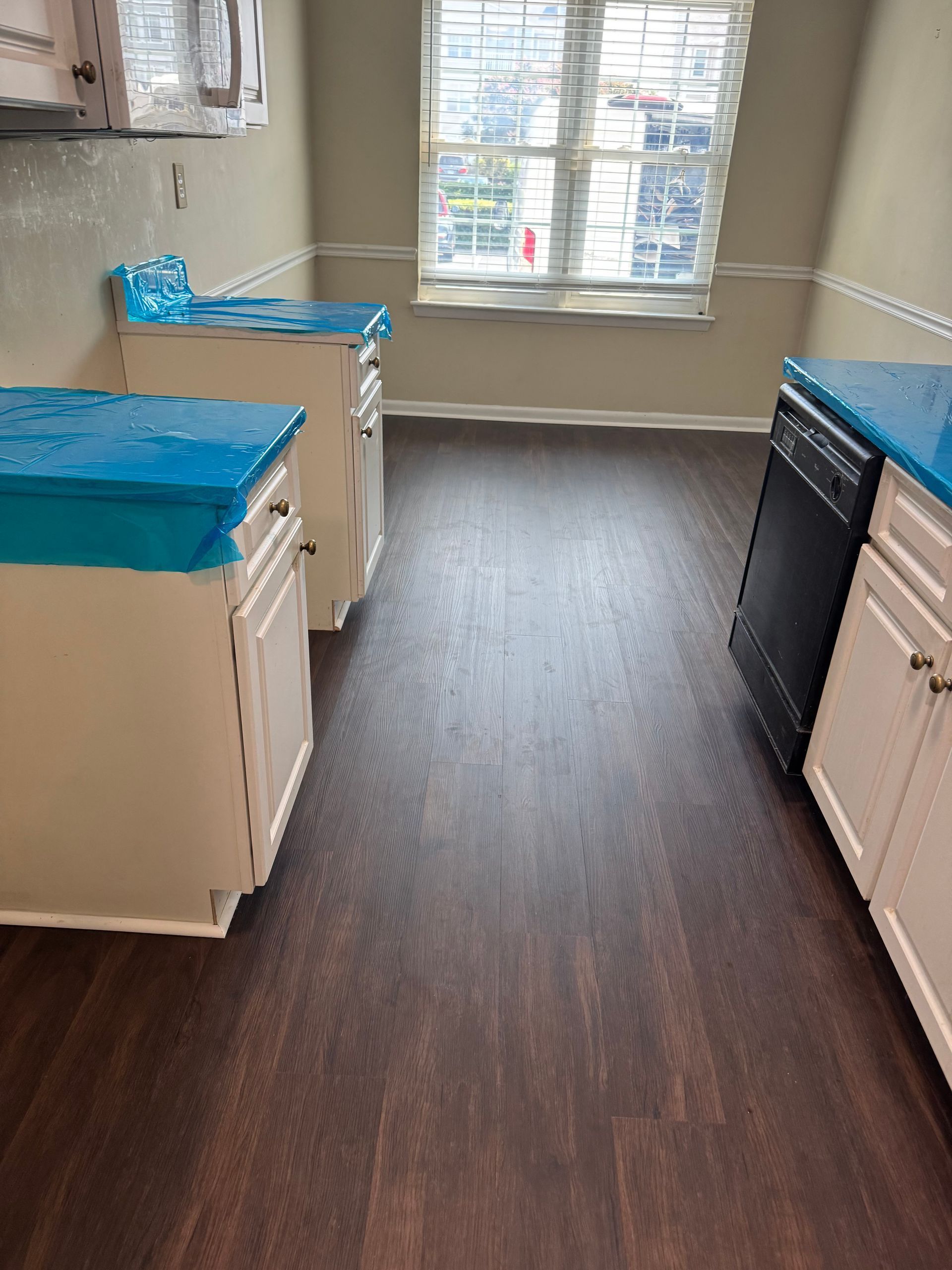 Empty kitchen with dark wood flooring, white cabinets, blue countertop covers, and a window at the end.
