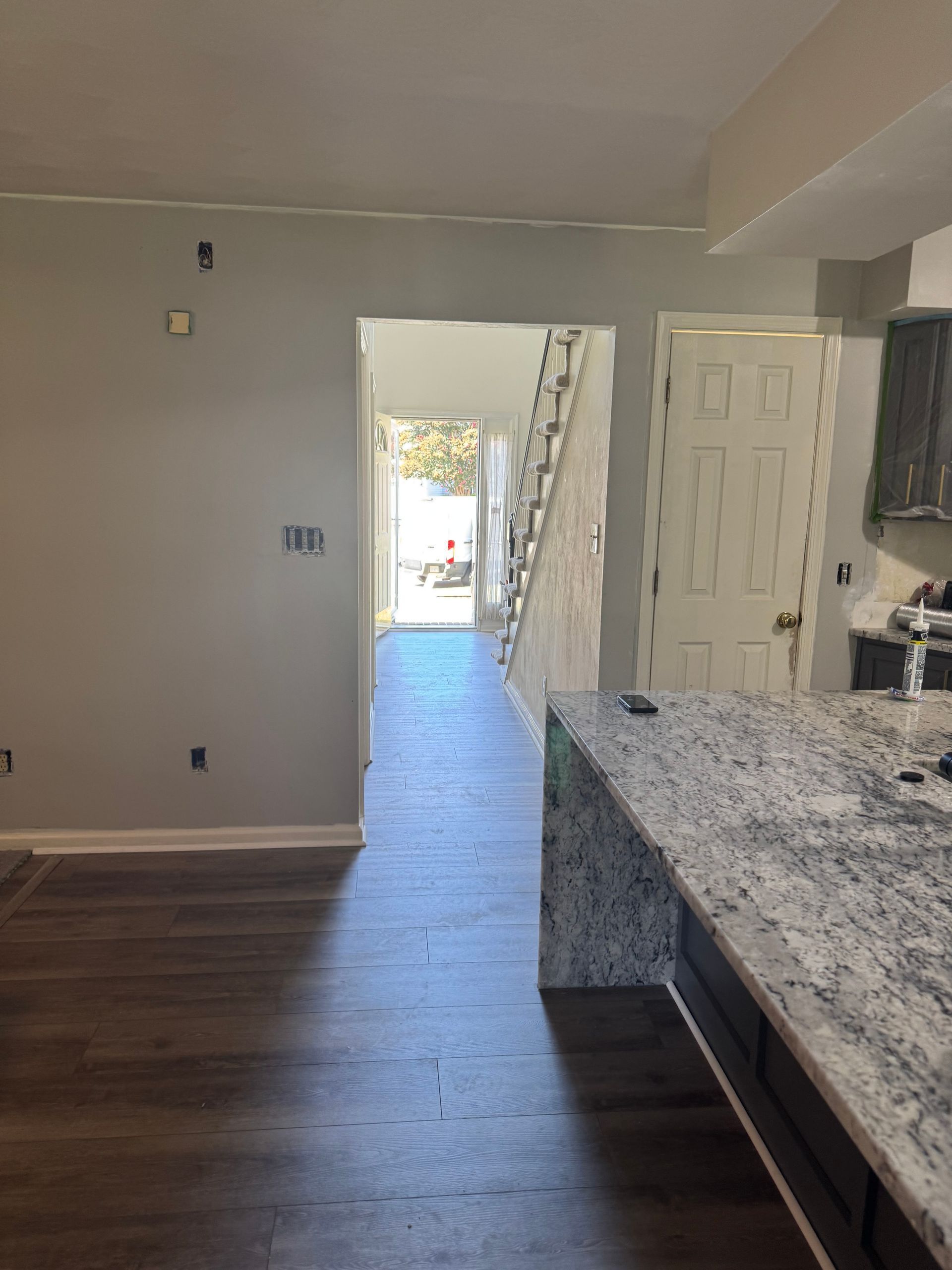 Empty modern kitchen with dark wood floor, white walls, granite island, and a bright hallway beyond.