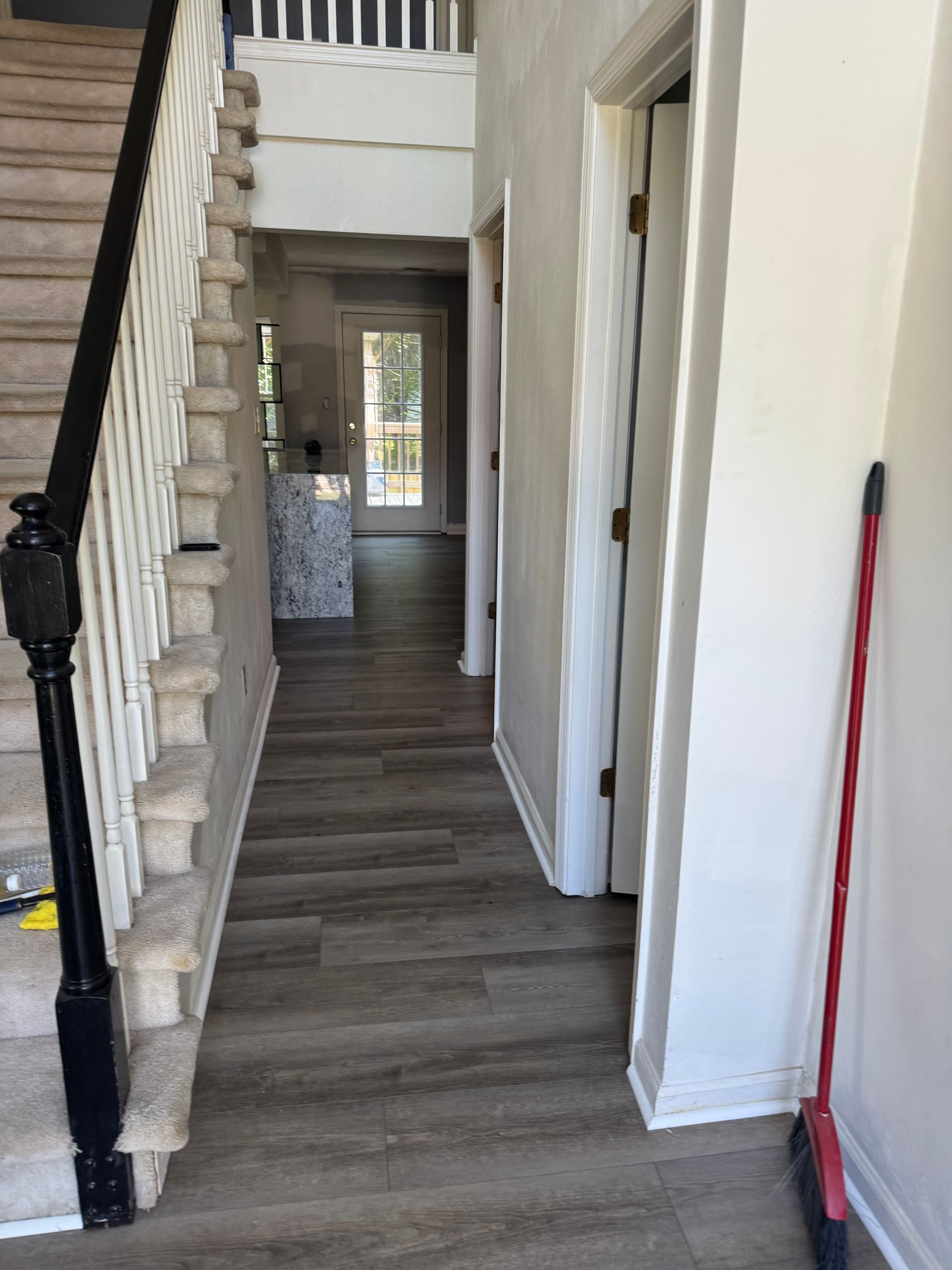 Narrow hallway with stairs on the left, white walls, gray wood floor, and a red broom leaning on the right.