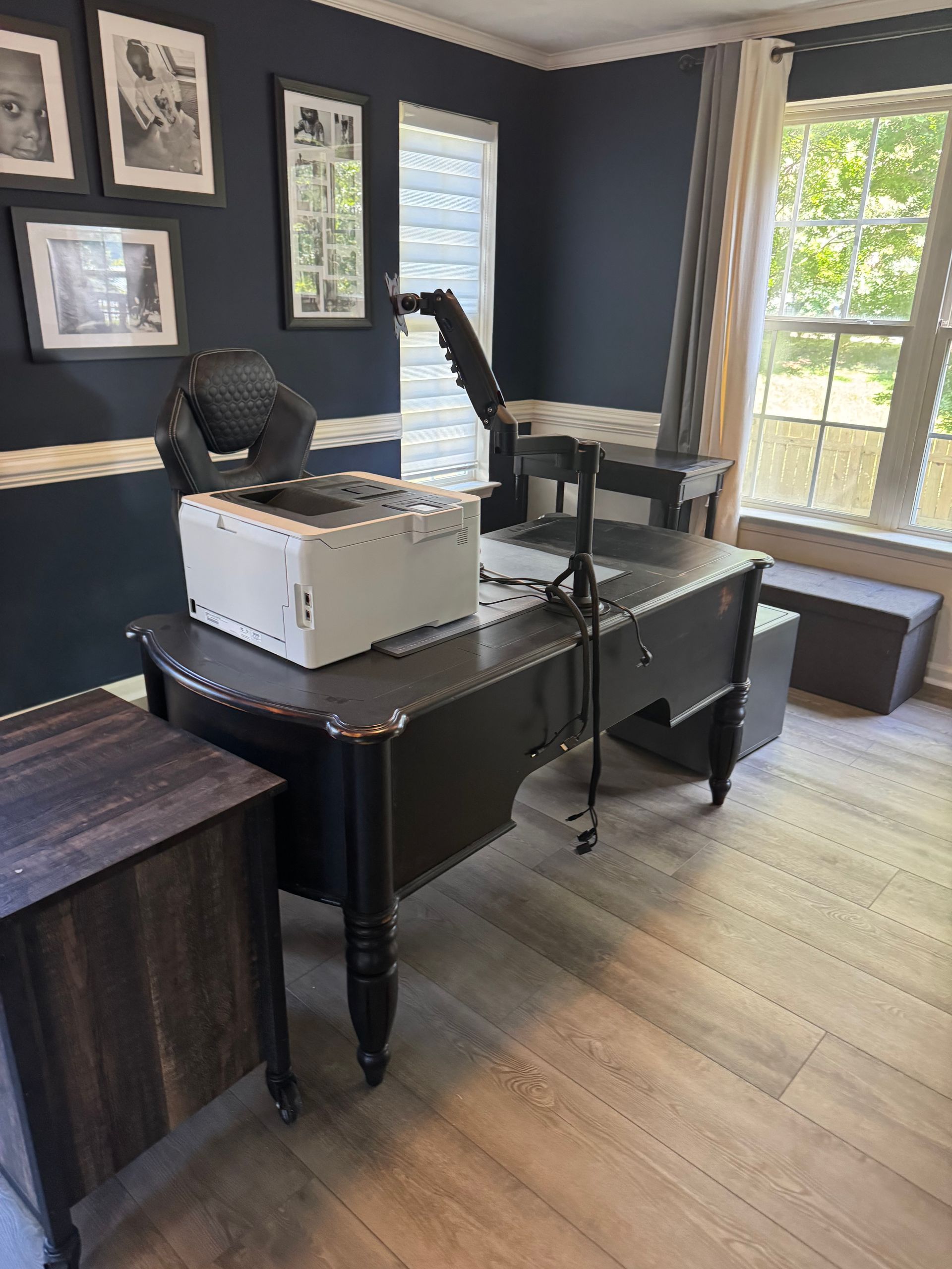 Home office with desk, chair, printer, and large windows in a dark-painted room.