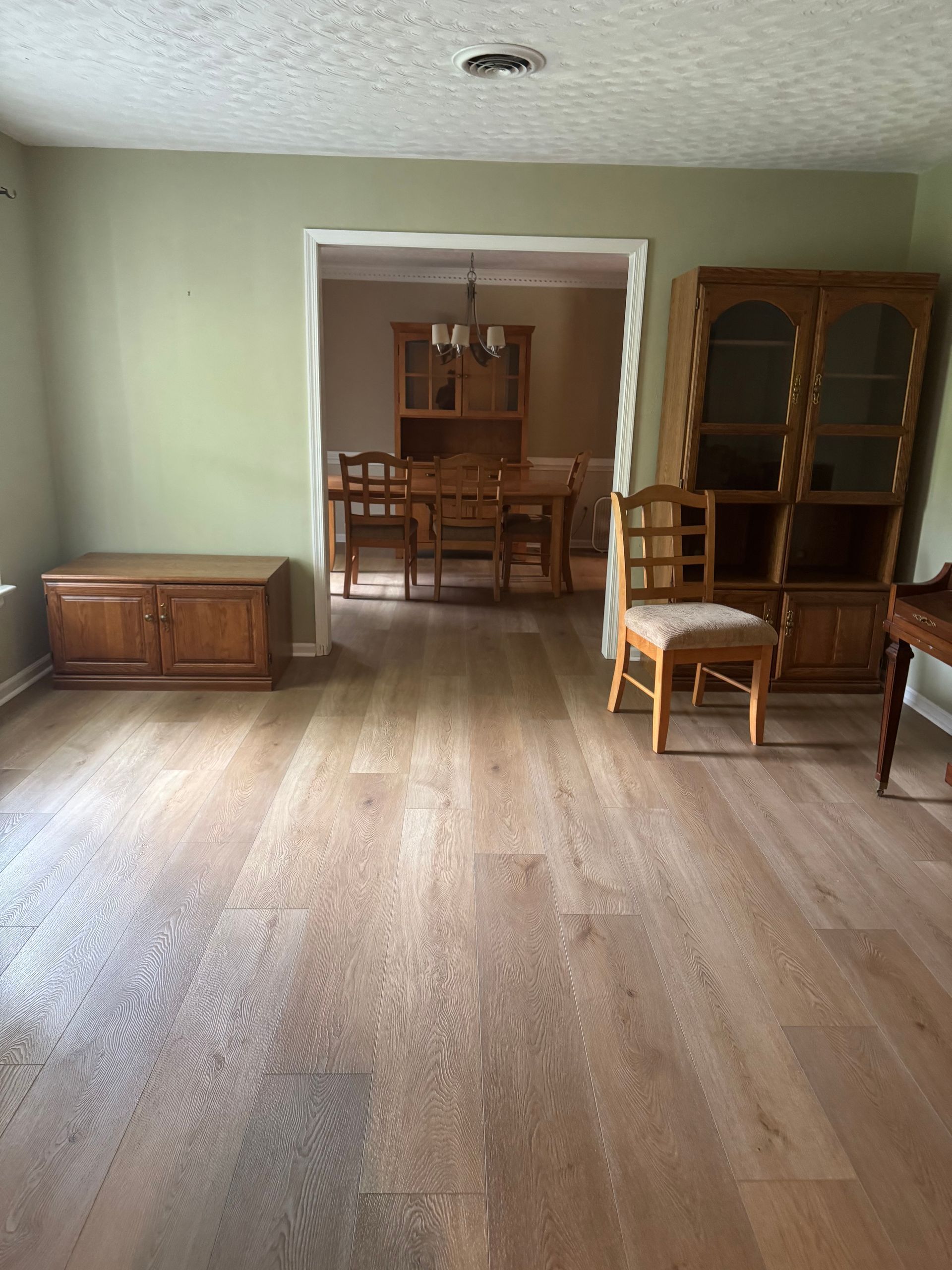 Empty living room with wood floor, pale green walls, and wooden cabinets opening to a dining area.