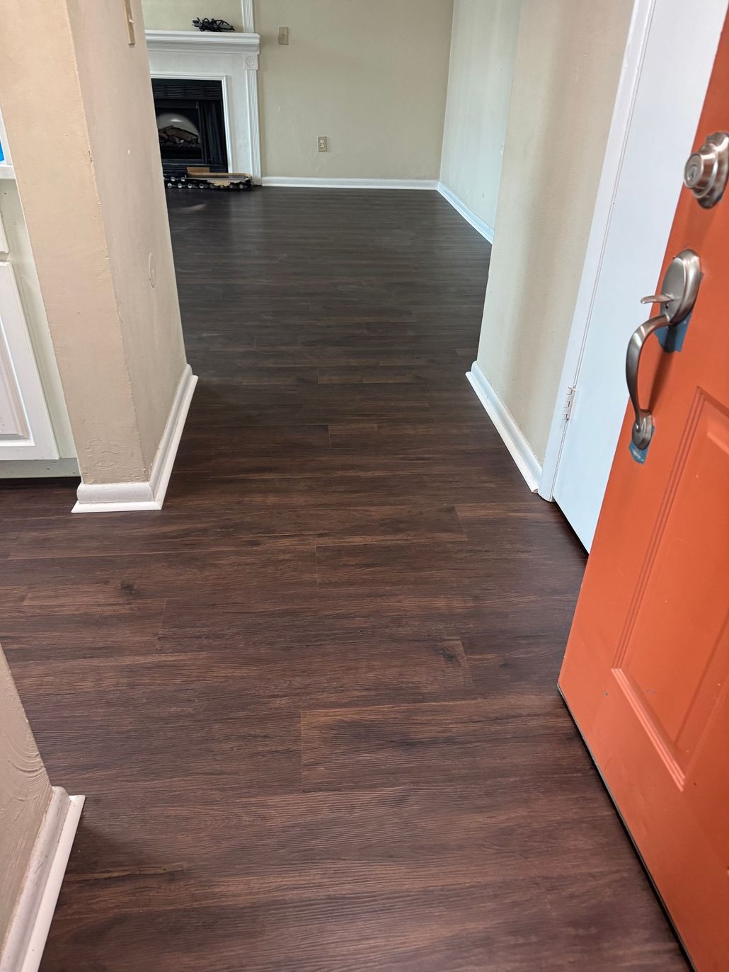 View through an open orange door into a hallway with dark wood flooring and a fireplace in the distance.