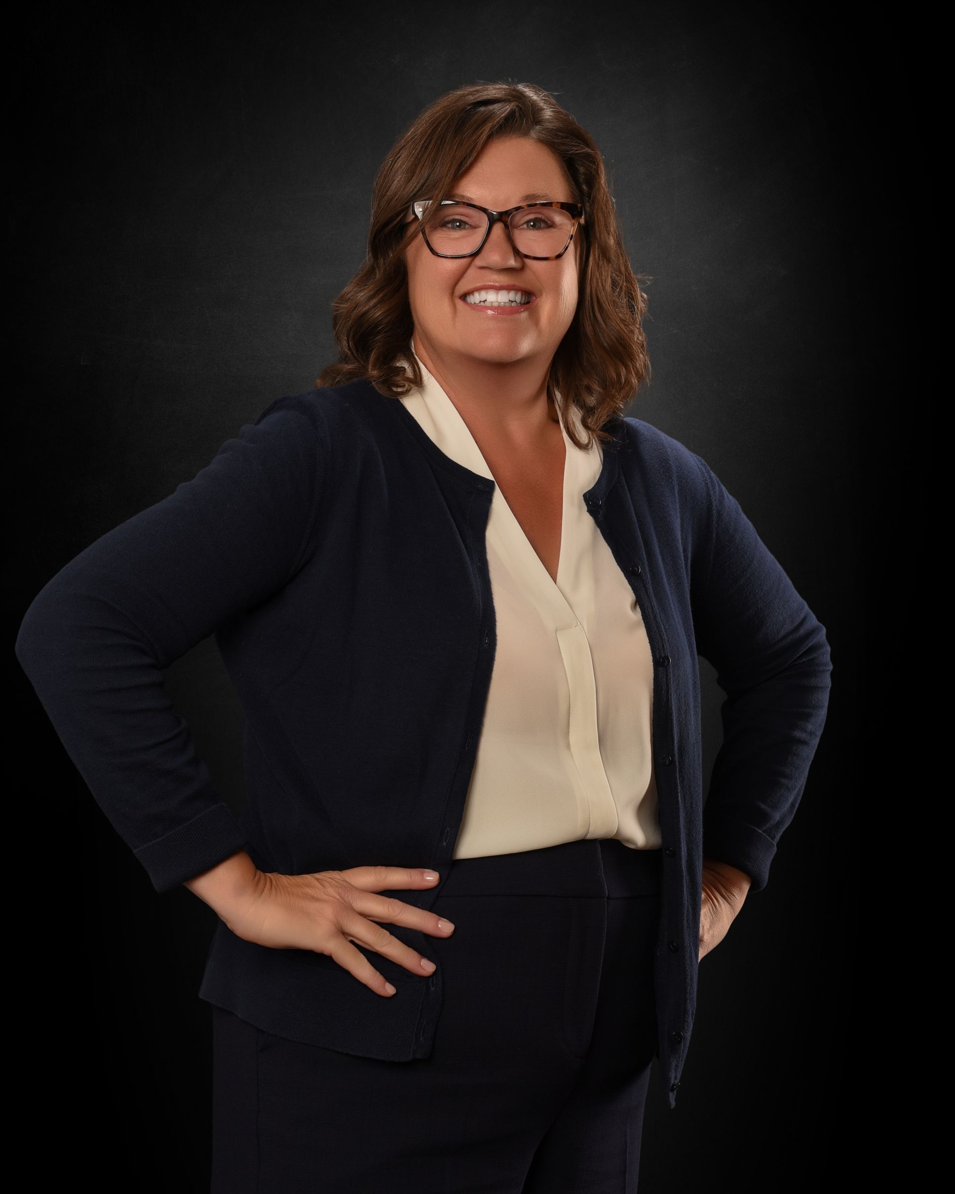 Woman standing with hands on hips, smiling in a navy cardigan and cream blouse against a dark background.