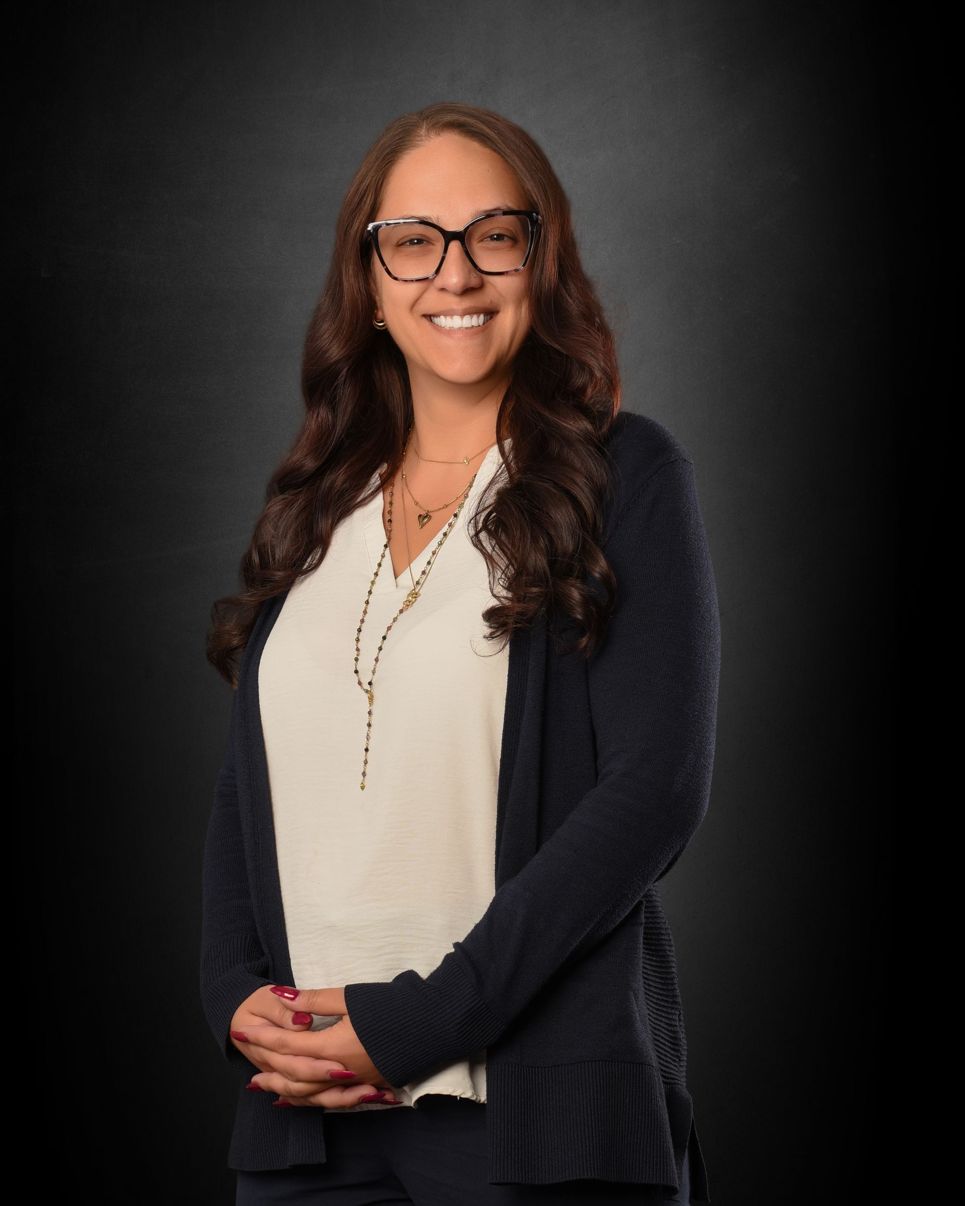 Smiling person with glasses in a cream blouse and dark cardigan, standing against a dark background.