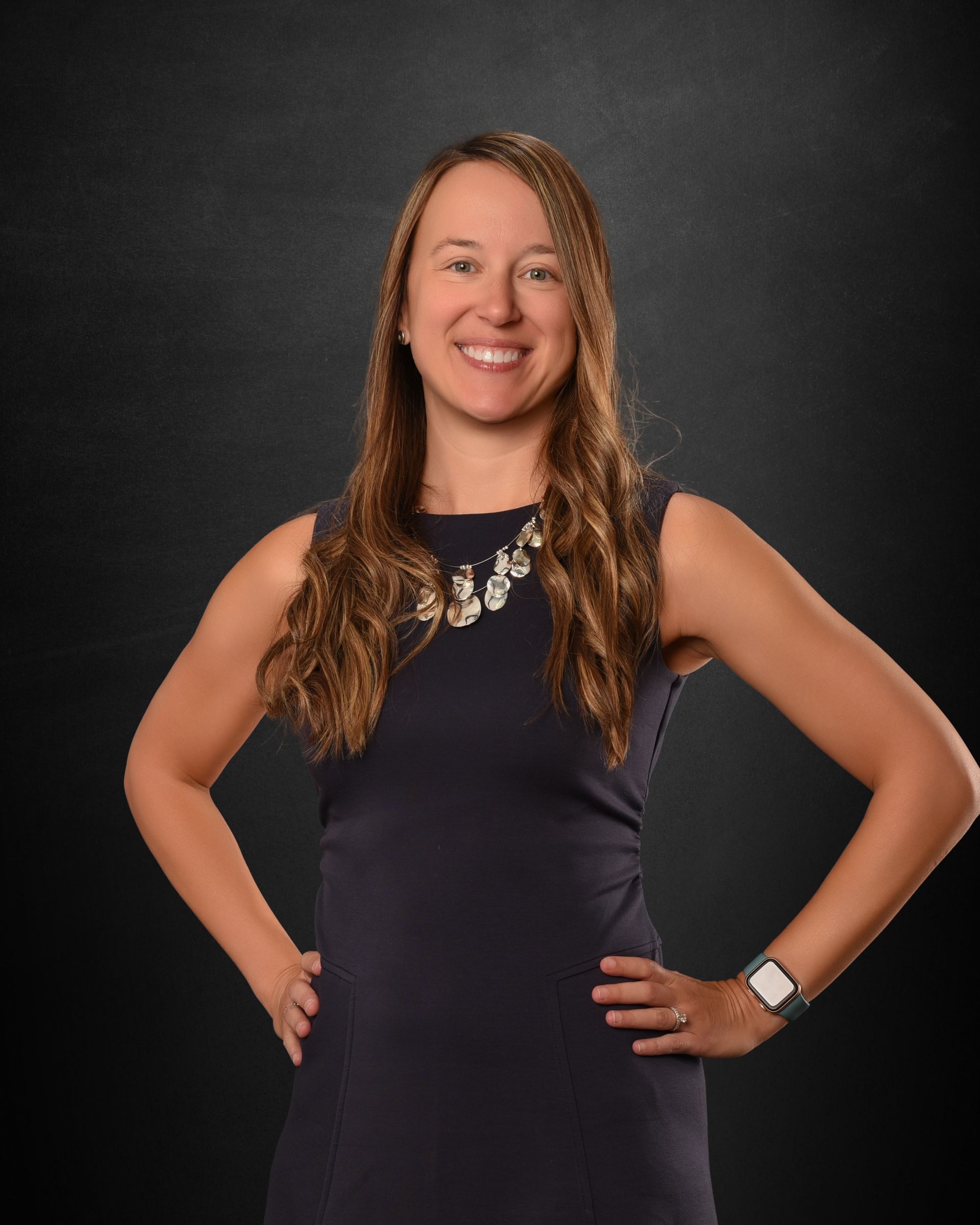 Smiling person in a sleeveless navy dress, hands on hips, against a dark studio backdrop.