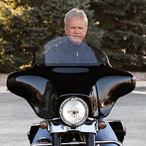 Man on a black motorcycle with a large windshield, riding on a road with trees in the background