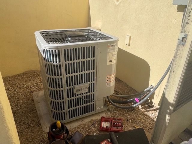 Gray air conditioning unit installed against a tan wall on a gravel bed; tools visible nearby.