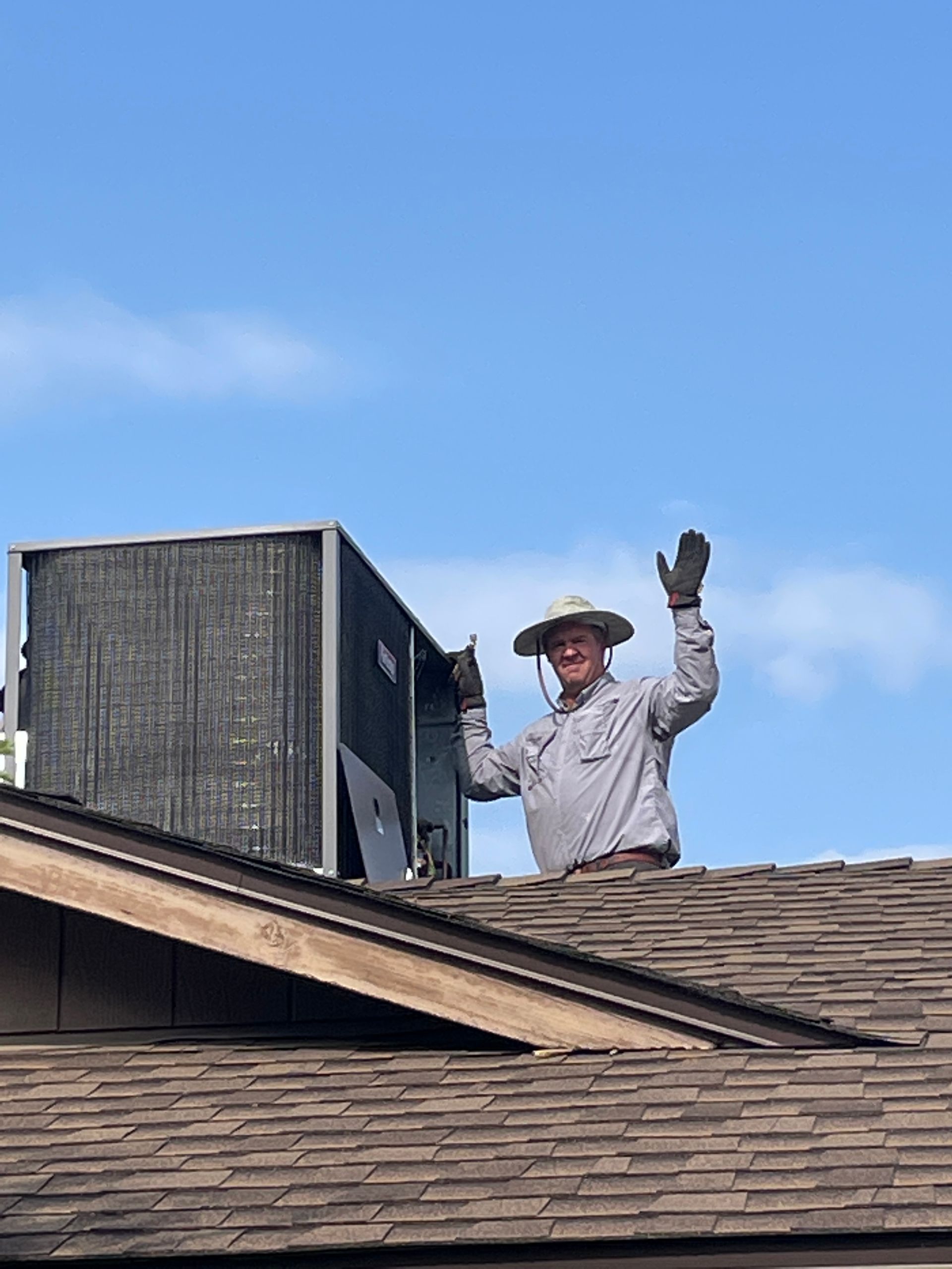 Man on rooftop waves, near HVAC unit. Blue sky, brown roof.