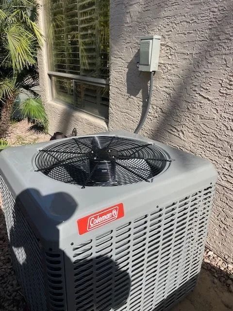 Coleman air conditioning unit outside near a window, connected to a power source on a stucco wall.