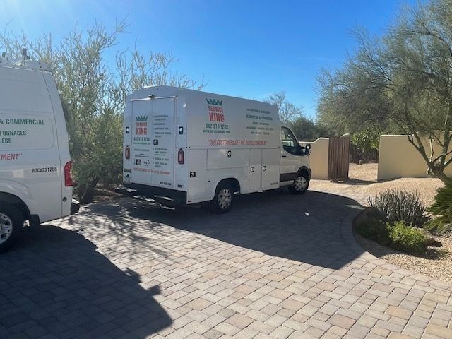 Two white service vans parked on a brick driveway near a house and desert vegetation under a sunny sky.
