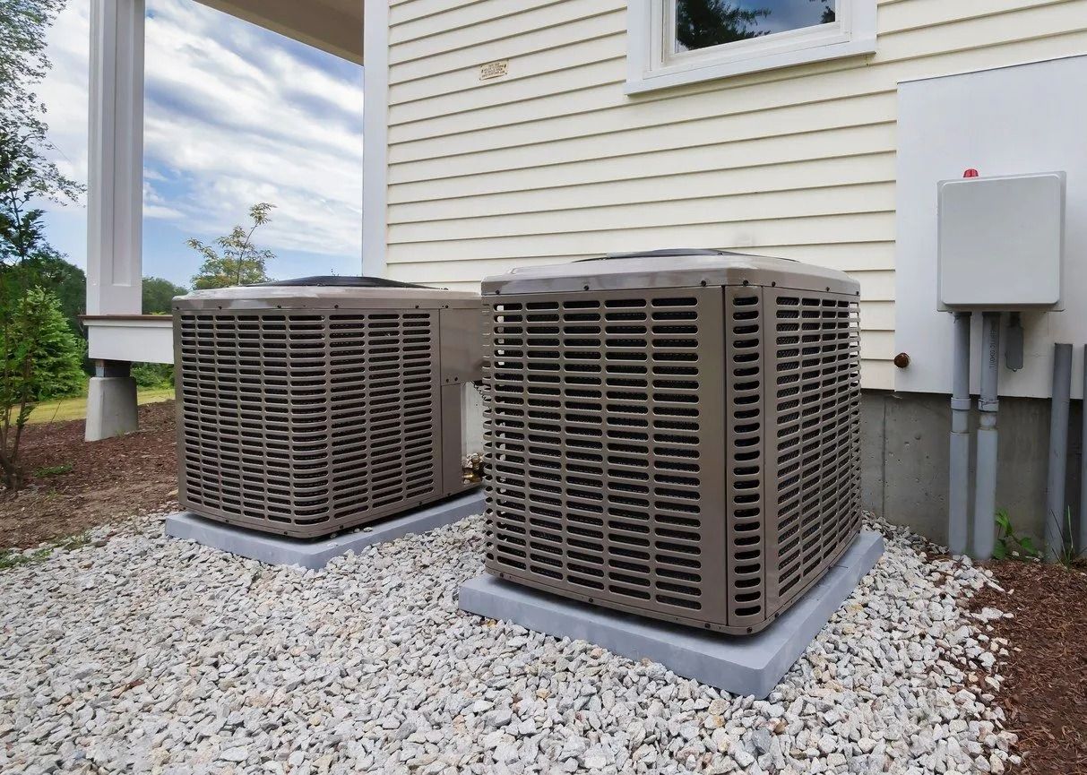 Two outdoor air conditioning units on gravel next to a house.