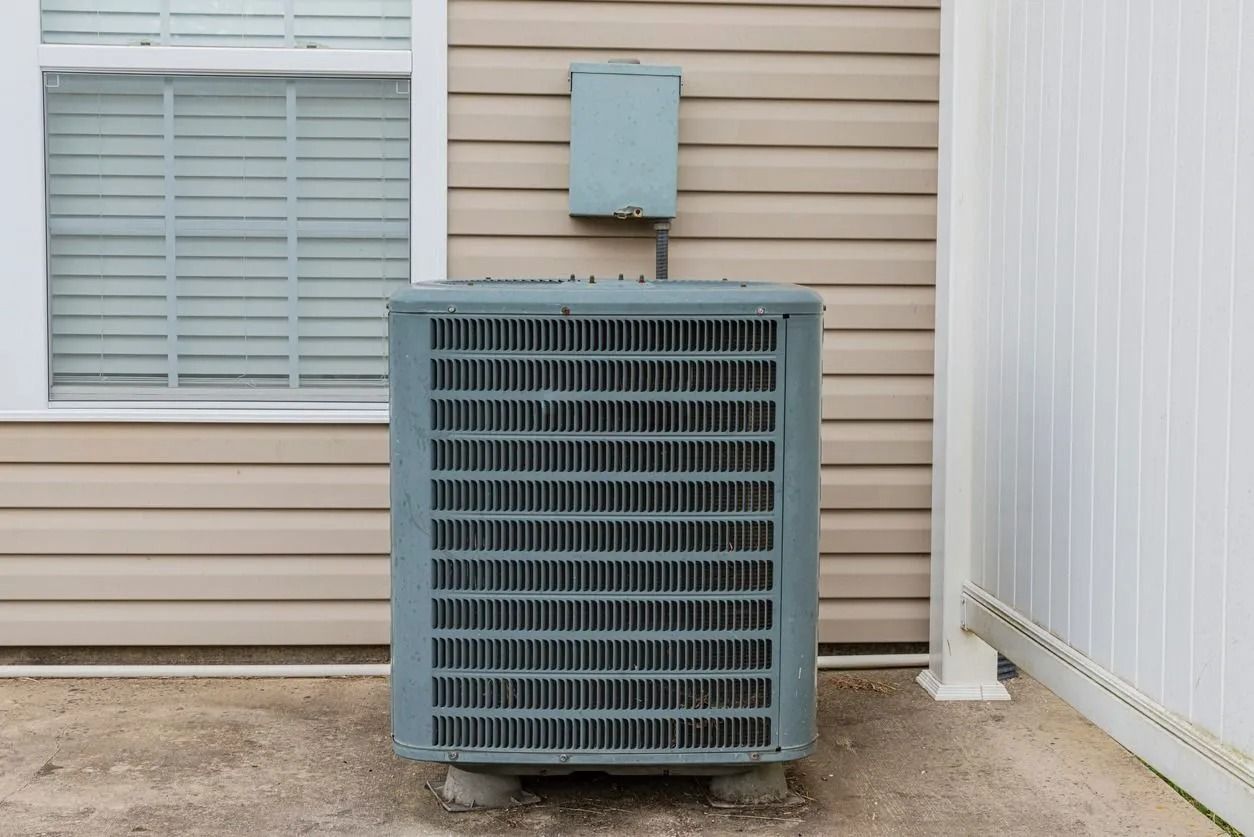 An air conditioning unit outside a building with beige siding and a window.