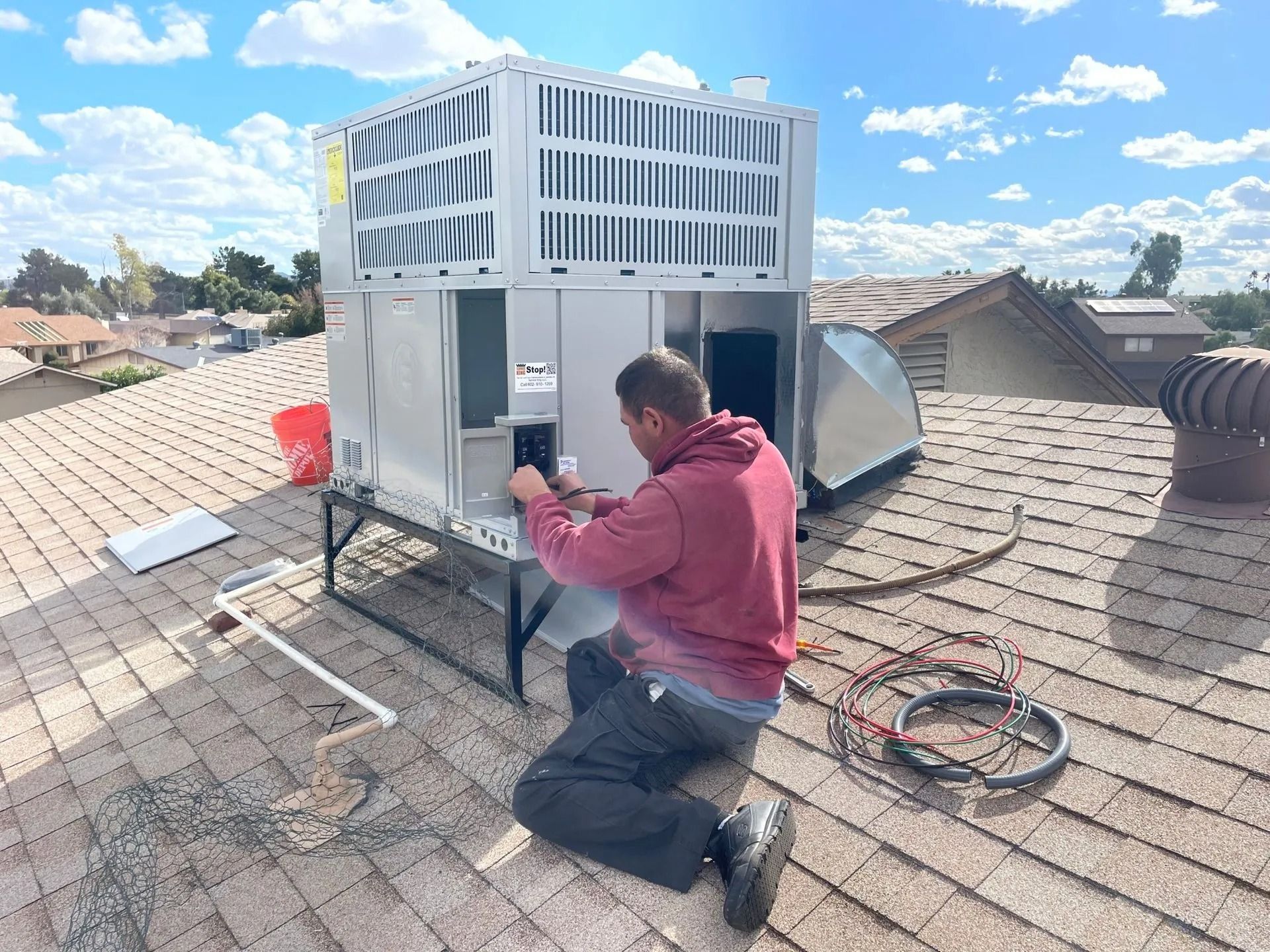 HVAC technician working on rooftop unit, kneeling on a brown shingled roof under a blue sky.