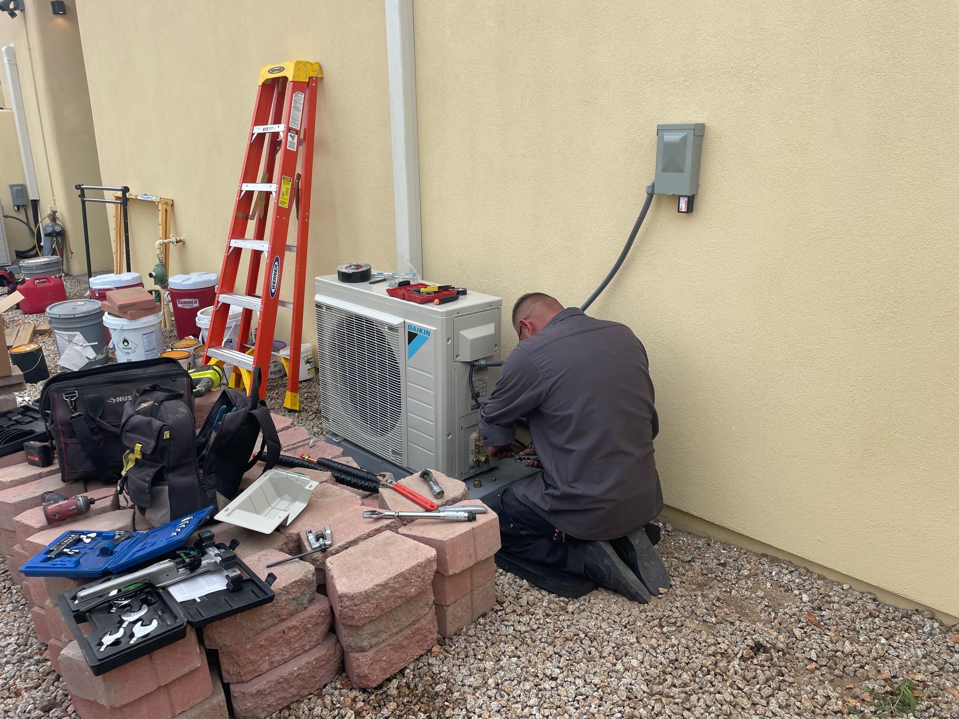 HVAC technician kneeling by an outdoor unit, working on it near a tan wall and tools.
