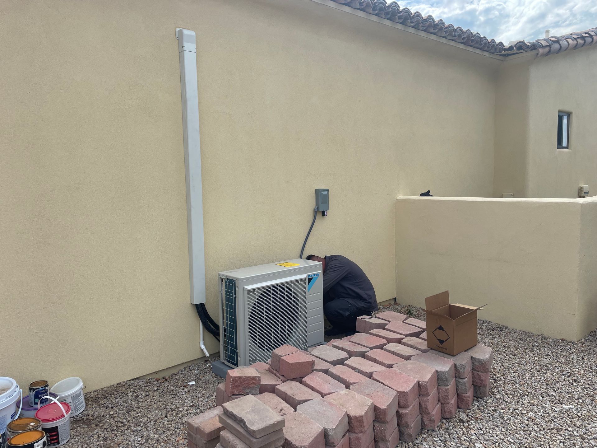 HVAC unit against a stucco wall, with brick pavers and a person working on it in a backyard setting.