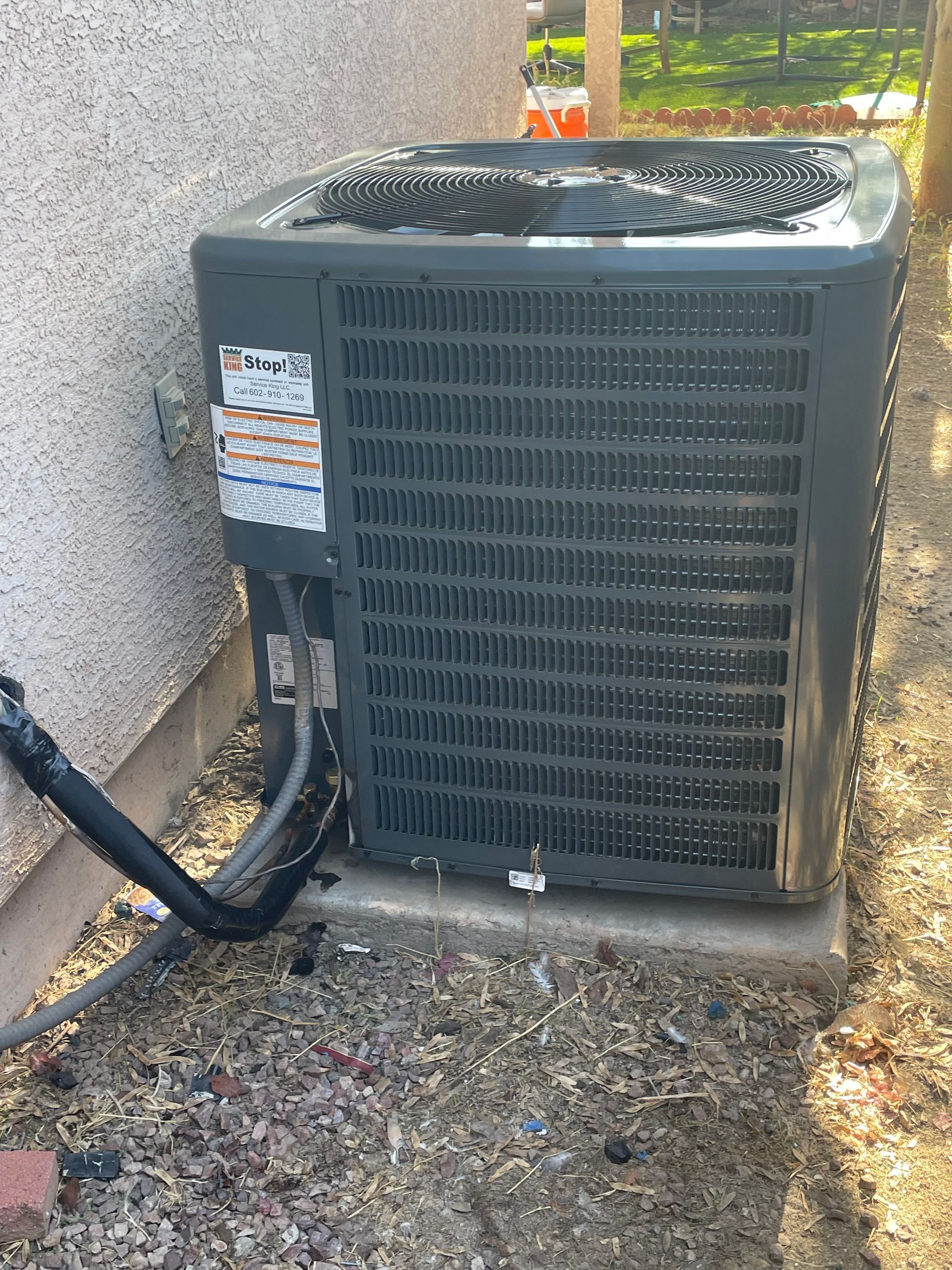 Exterior air conditioning unit next to a stucco wall, surrounded by dry grass and debris.