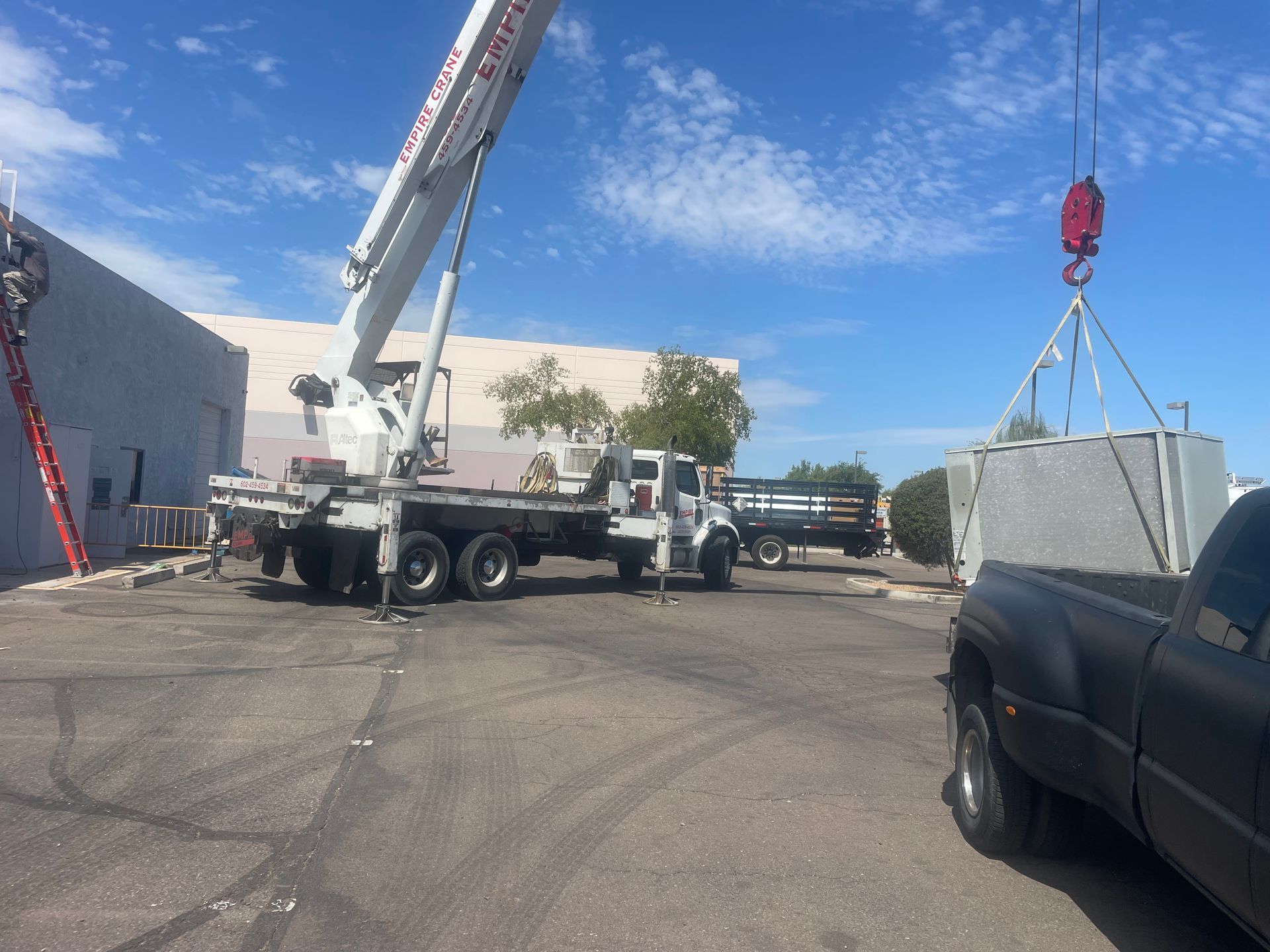 Crane lifting a large gray object from a flatbed truck near a building; sunny day.