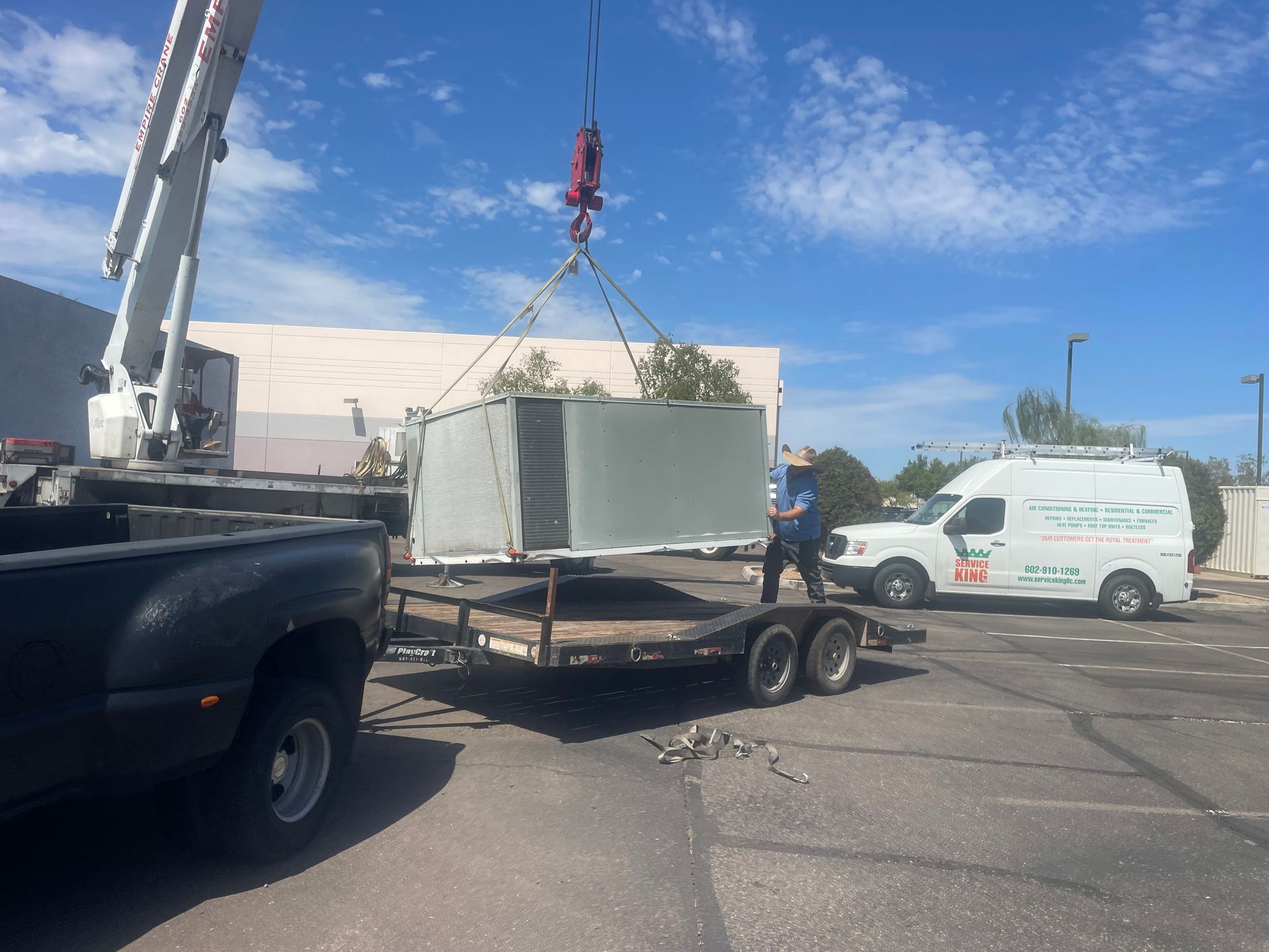 A crane lifts an HVAC unit off a trailer. A worker stands nearby as a white van sits in the parking lot.