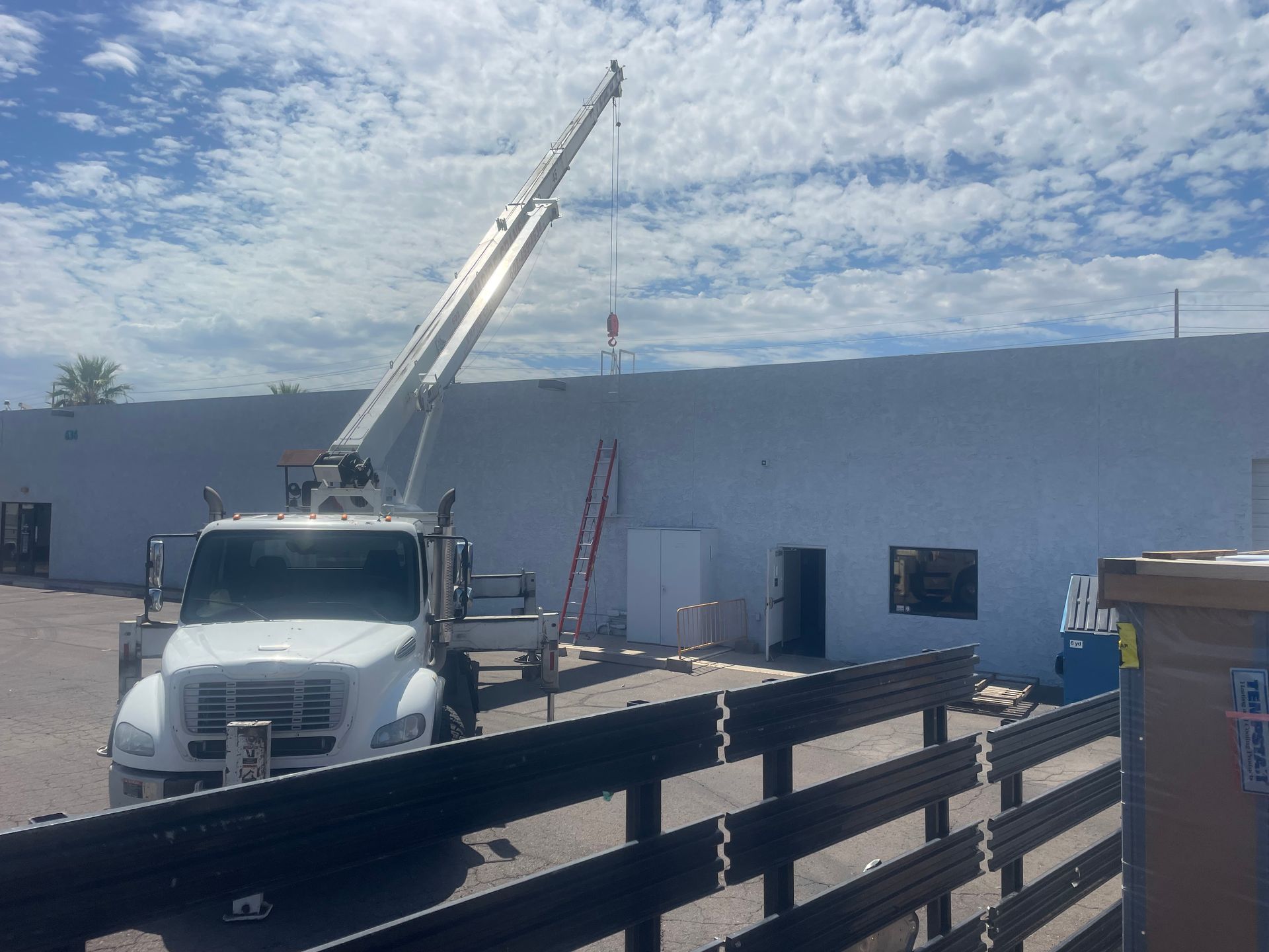Crane lifting something on a white building wall. Cloudy sky. Truck and ladder in view.