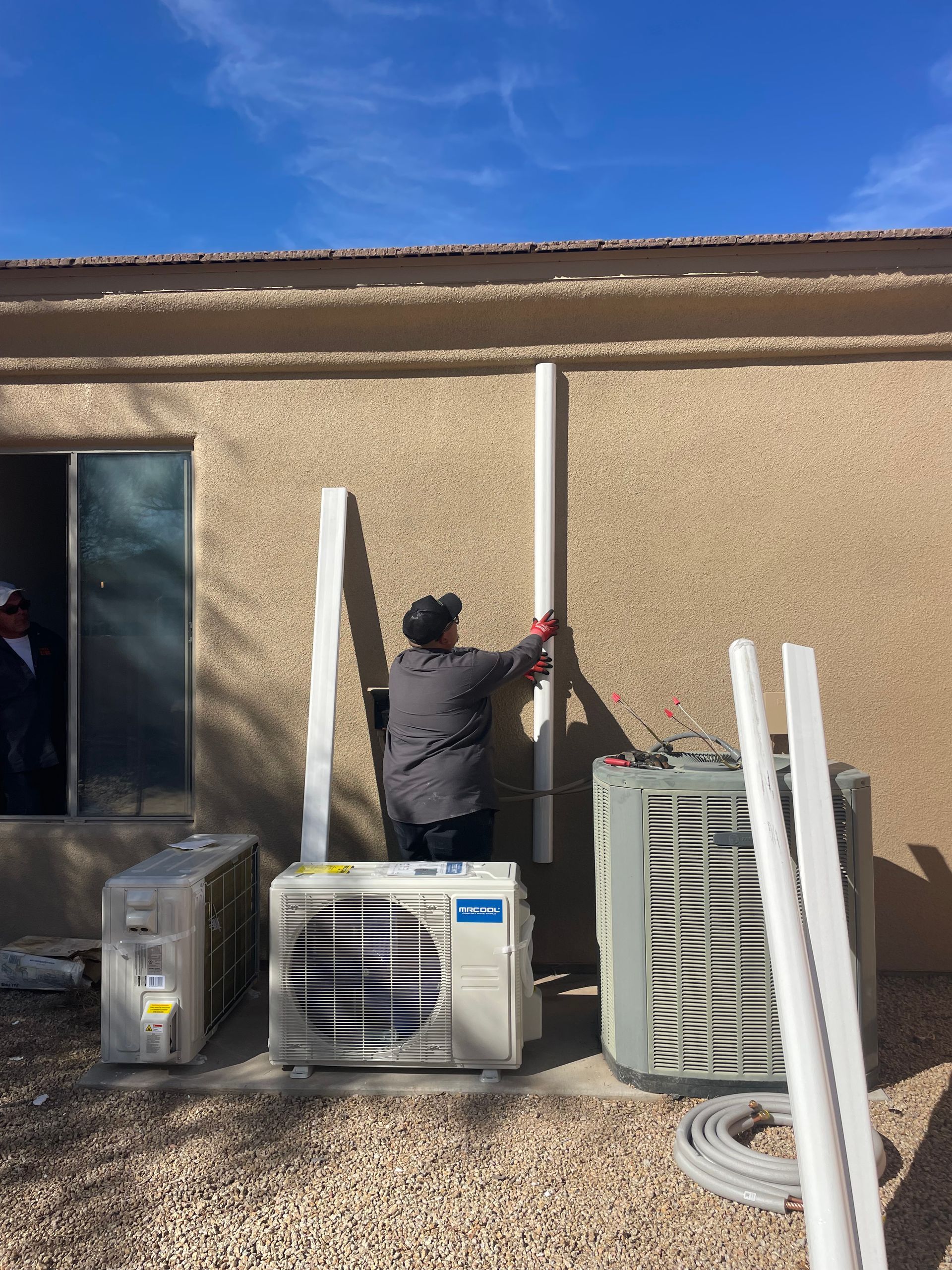 Person installs white PVC piping on a stucco wall outside, next to HVAC units and spare parts.
