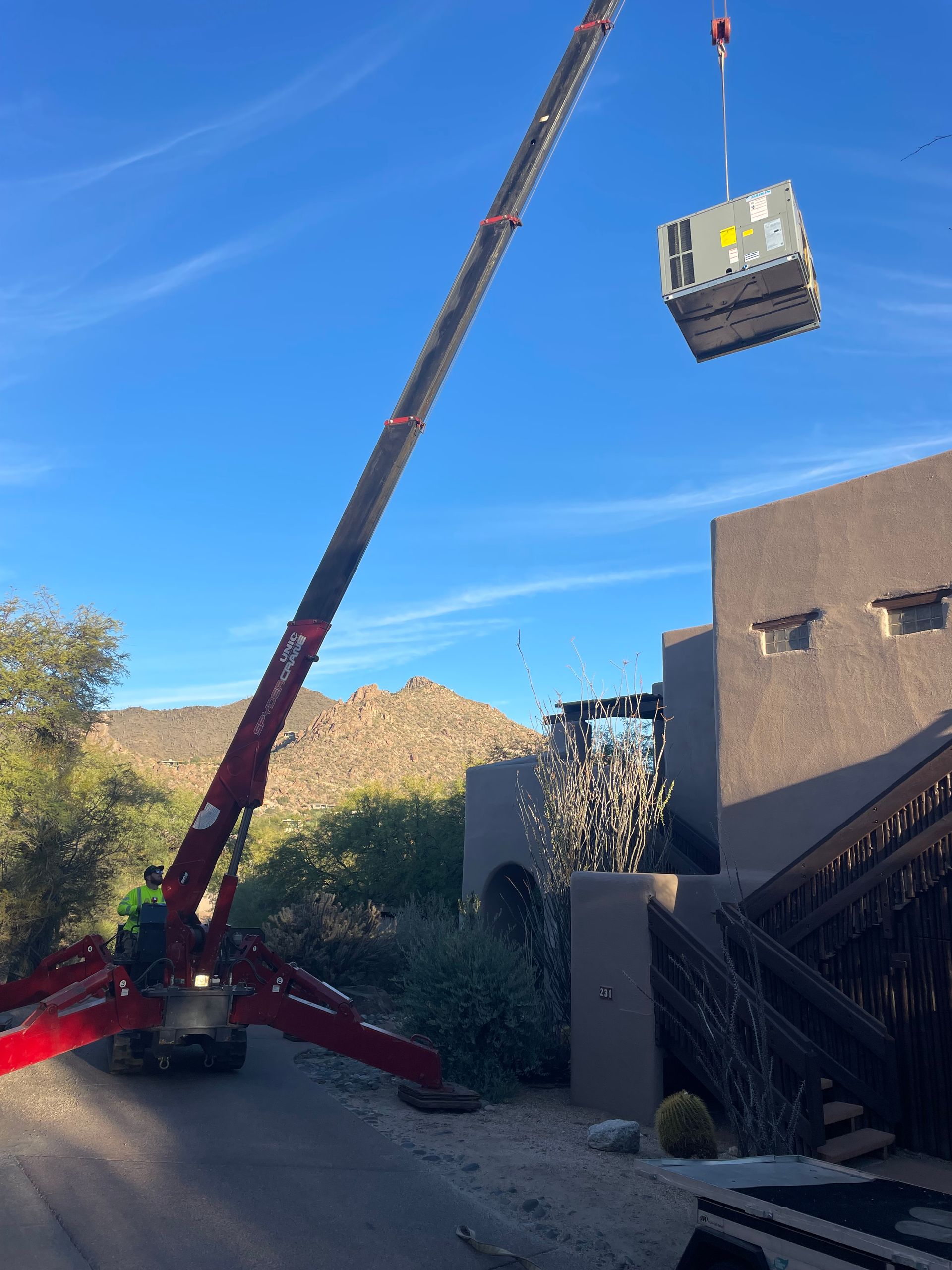 A crane lifts an HVAC unit towards a house against a blue sky, mountain in background.