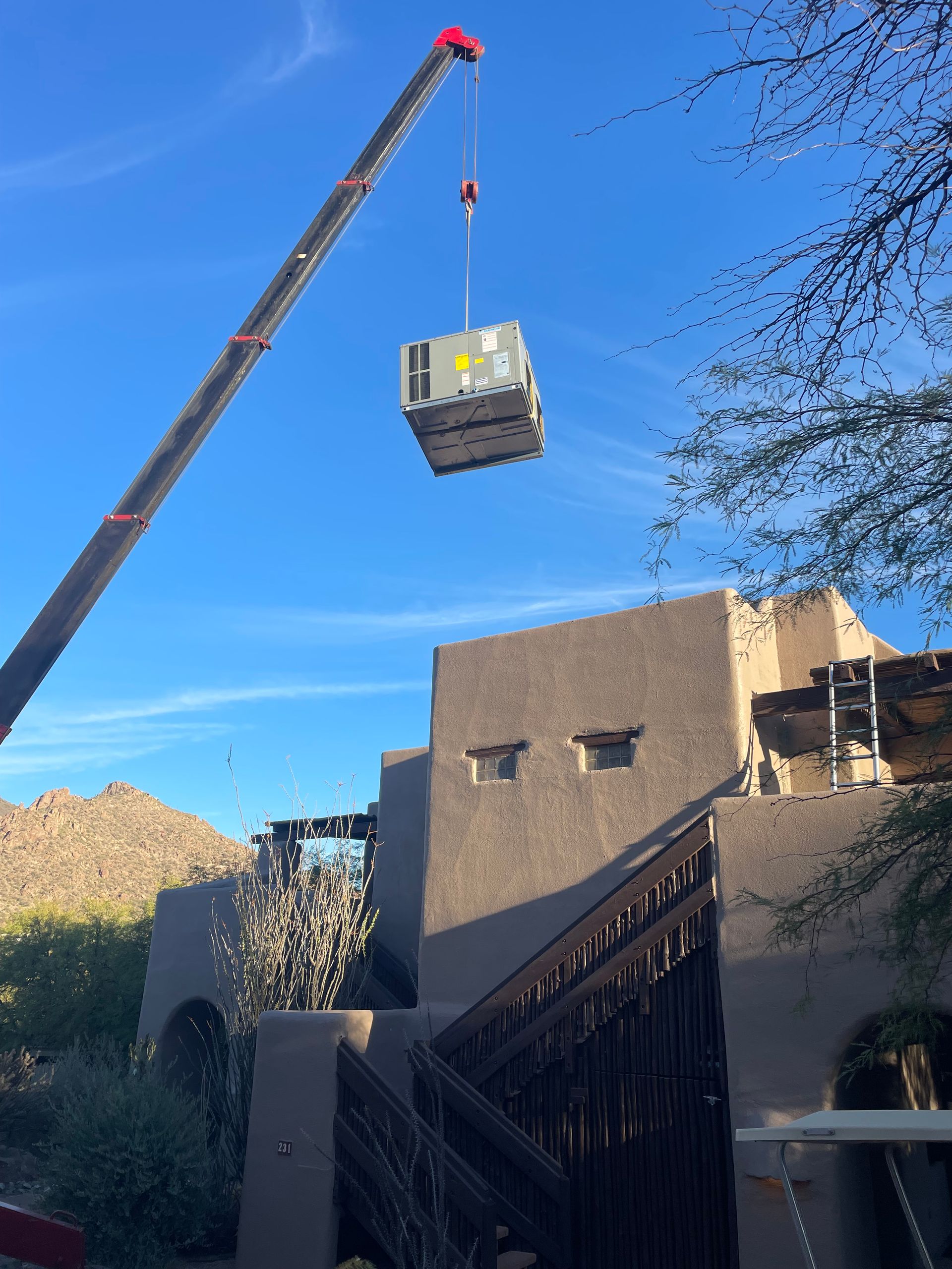 Crane lifting an HVAC unit over a two-story adobe-style building on a sunny day.