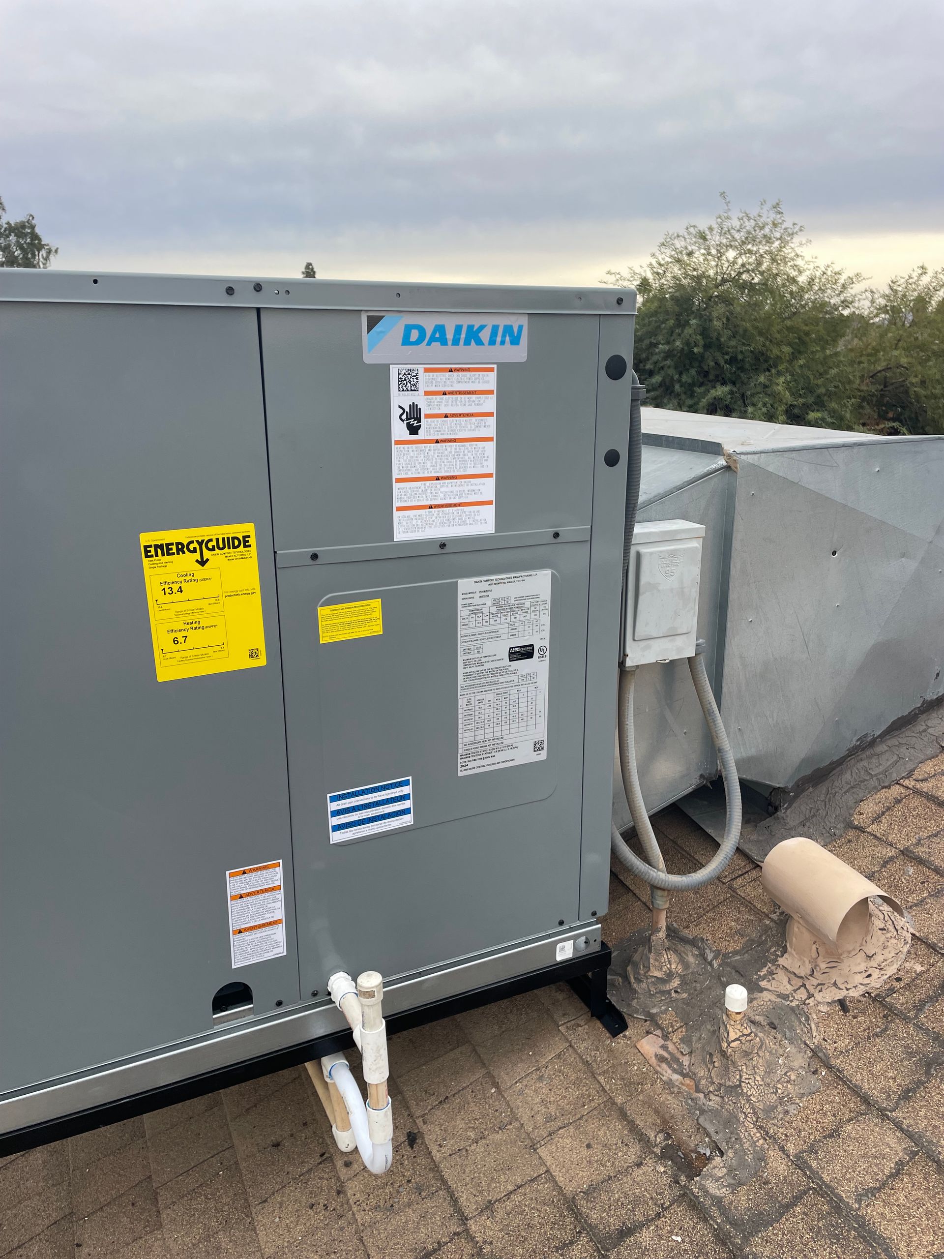 Daikin HVAC unit on a rooftop, with ductwork and various stickers, under an overcast sky.