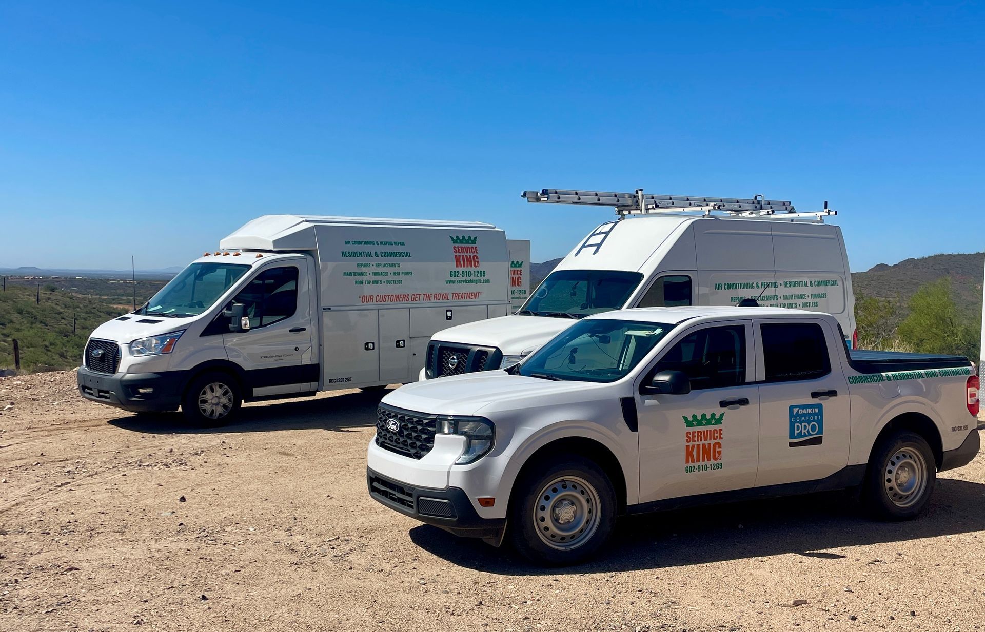 Three white work vehicles parked on a dirt road: a truck and two vans under a blue sky.