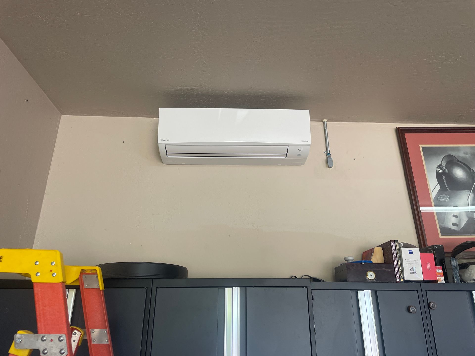 White air conditioning unit mounted on a beige wall above dark cabinets and a red ladder.