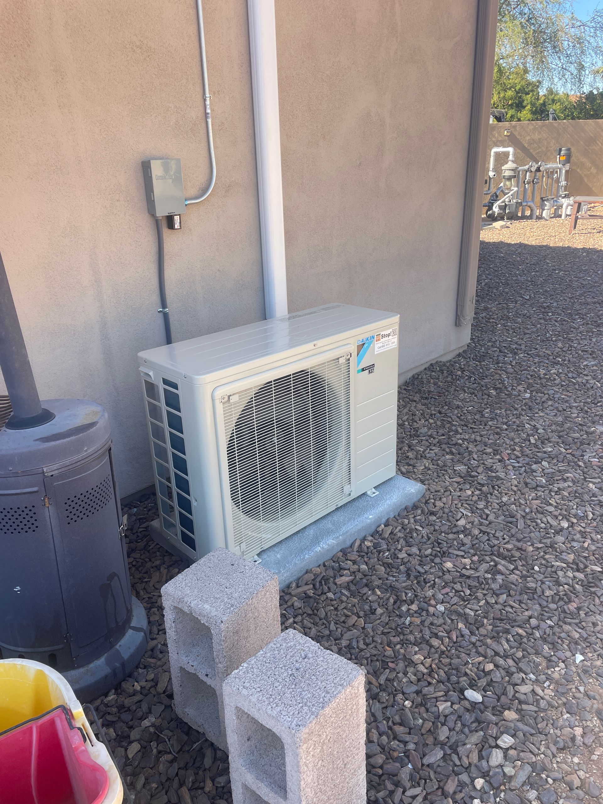 Exterior AC unit against a tan stucco wall. Two cinder blocks are in front of it on gravel.