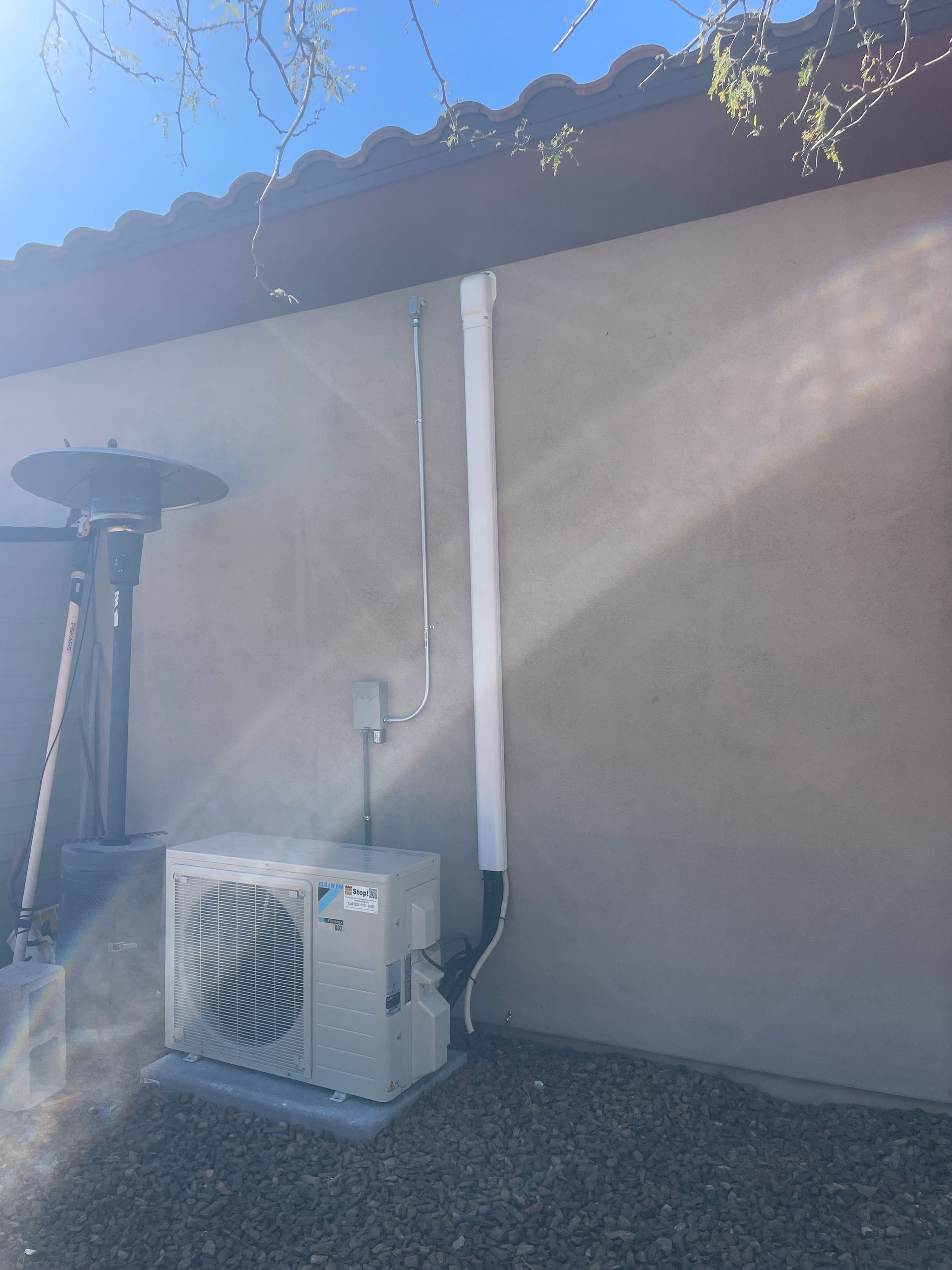Outdoor air conditioning unit against a stucco wall, with insulated pipes and a patio heater nearby.