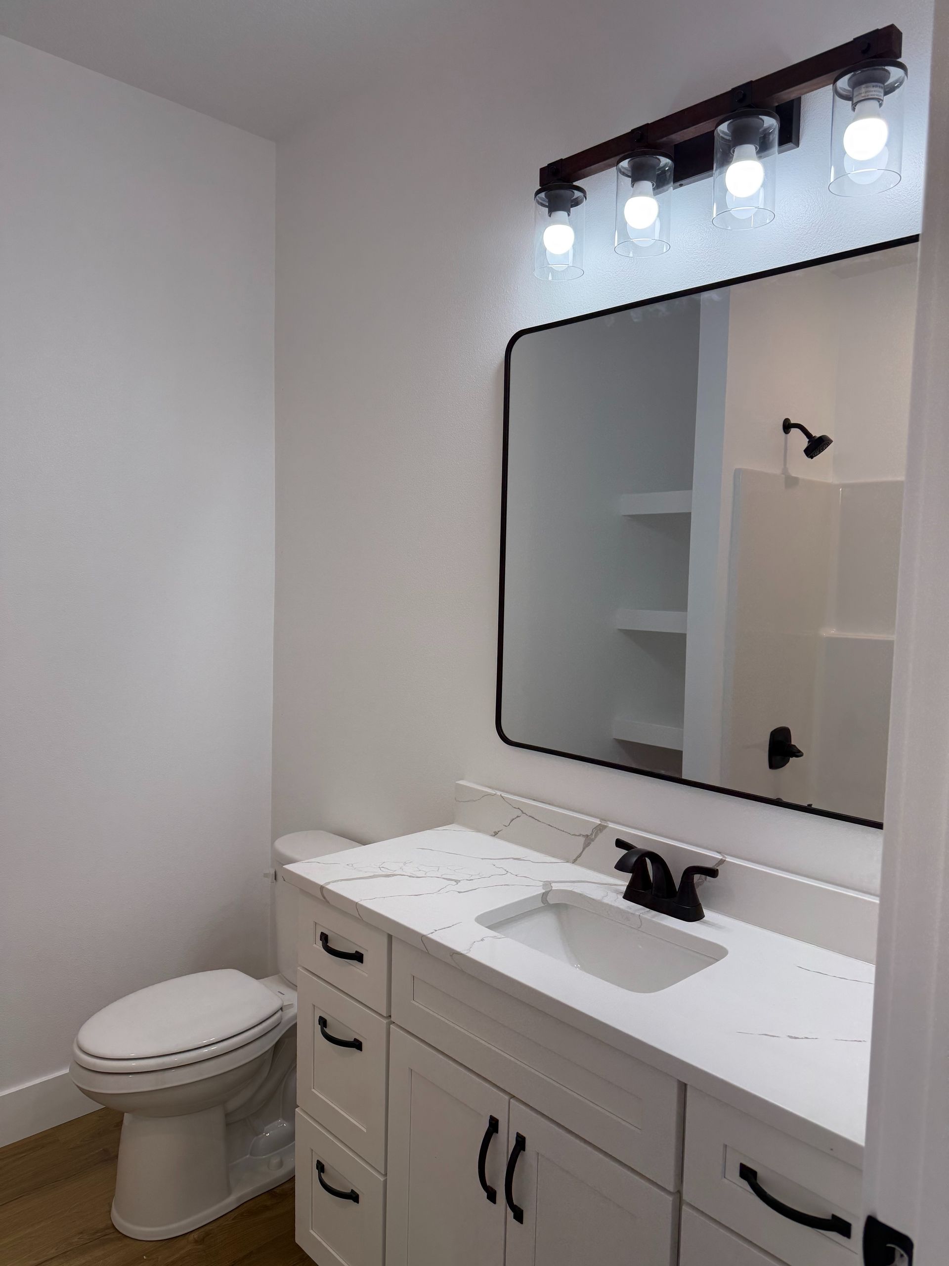 Small white bathroom with toilet, vanity sink, mirror, and lit light fixture above.