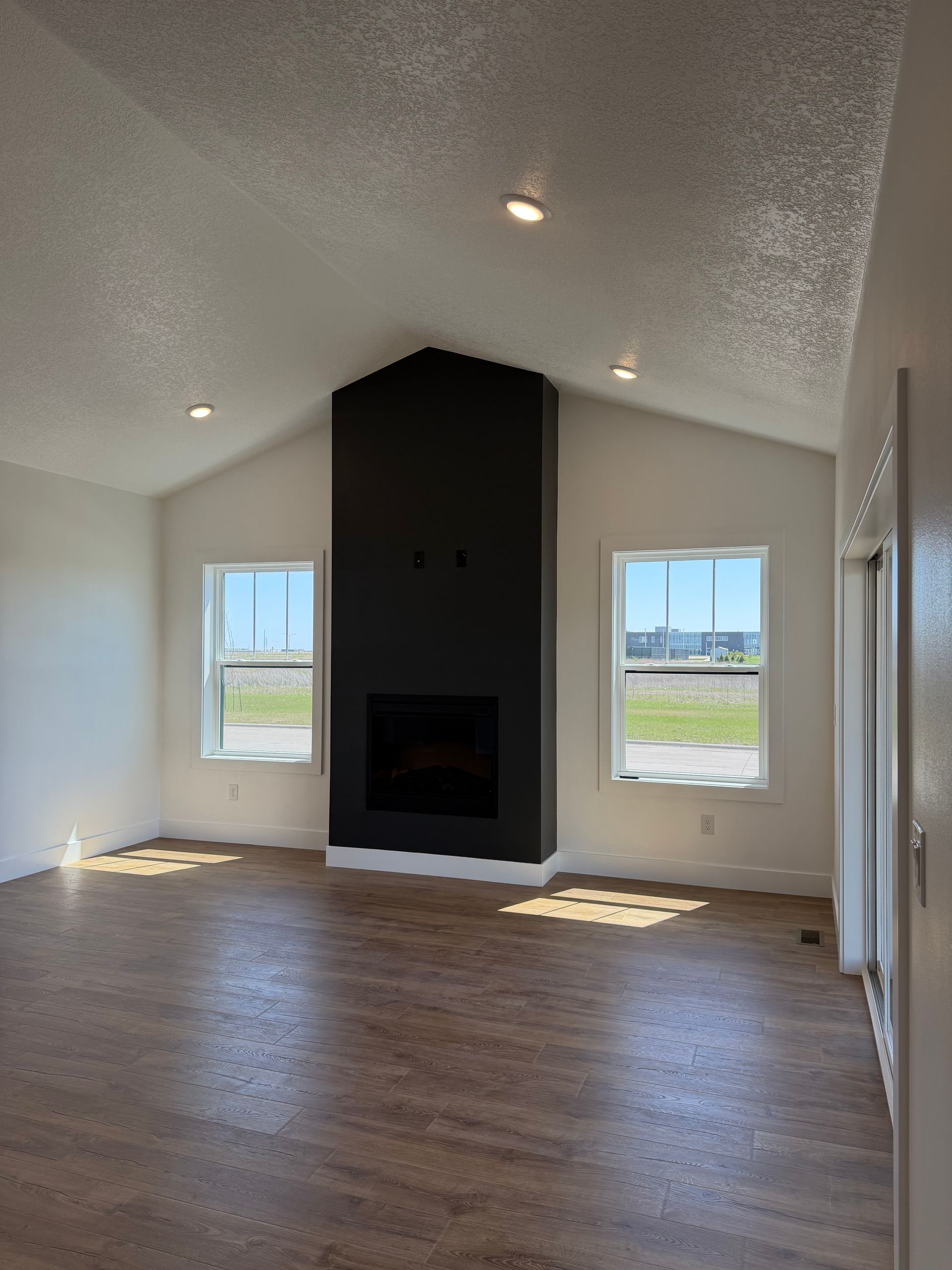 Empty living room with wood floors, white walls, black fireplace, and two windows letting in sunlight