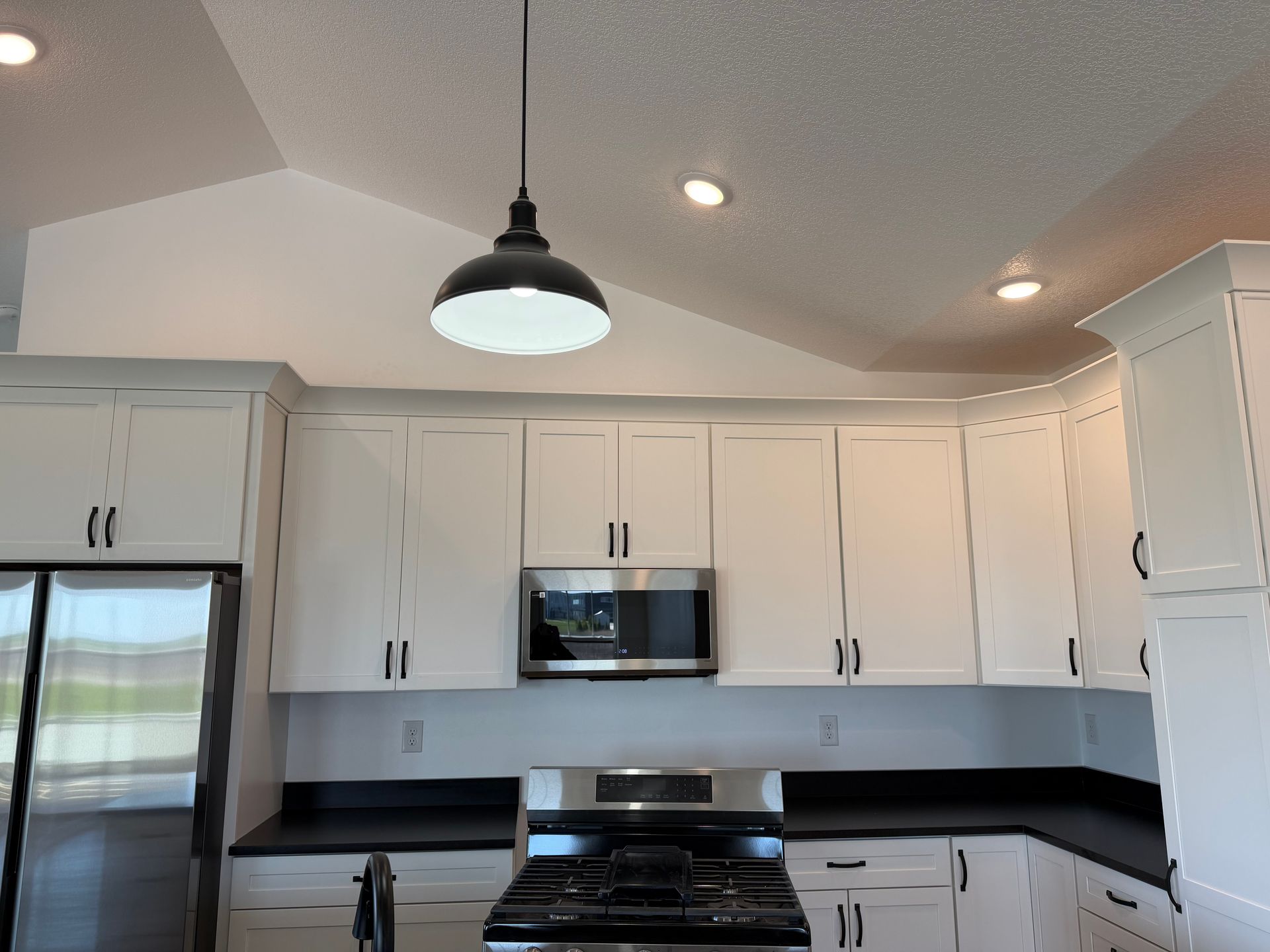 Bright white kitchen with black countertops, stainless steel appliances, and a vaulted ceiling light.