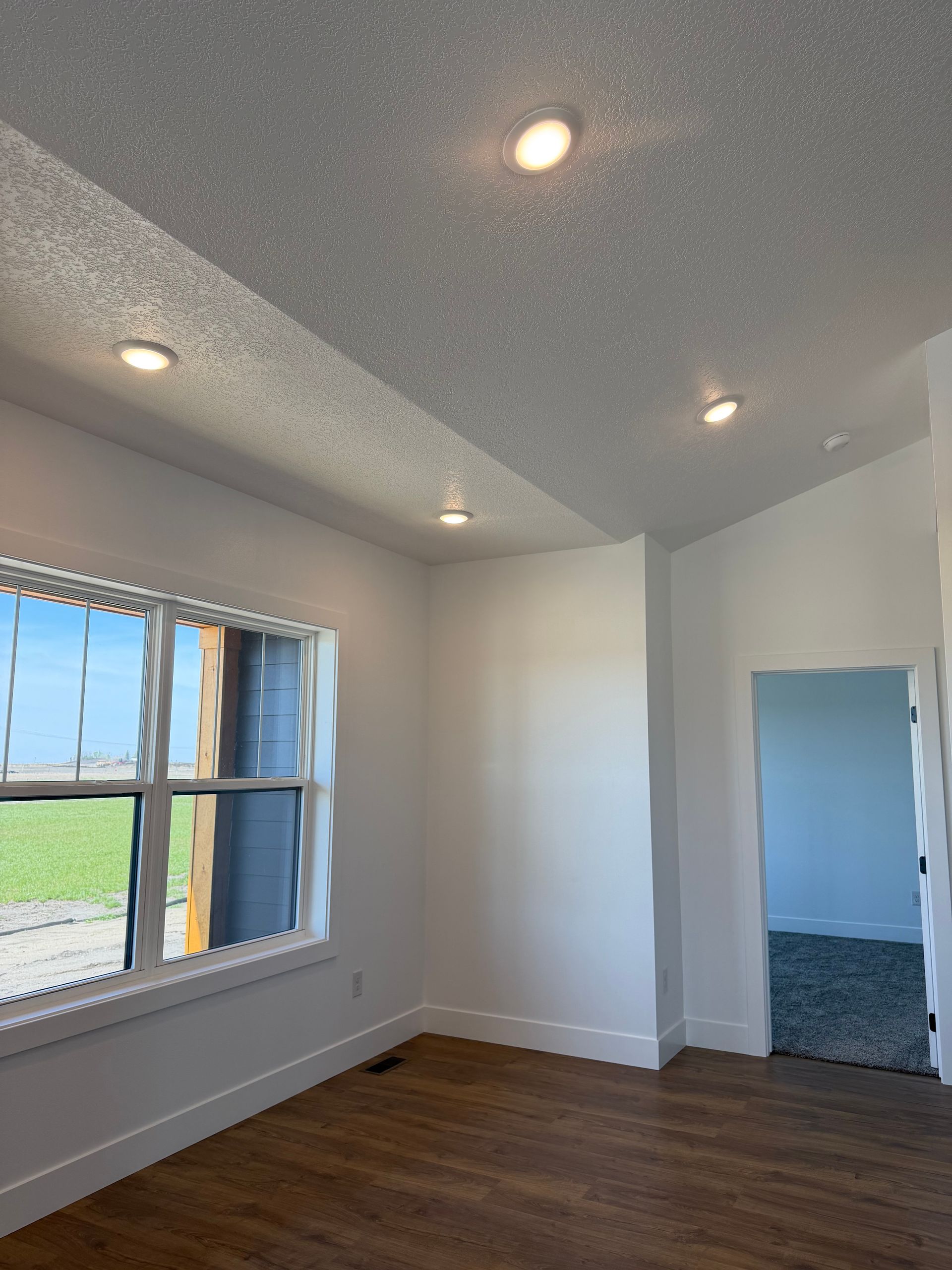 Empty white room with recessed lights, wood floor, and a window looking out to grass.