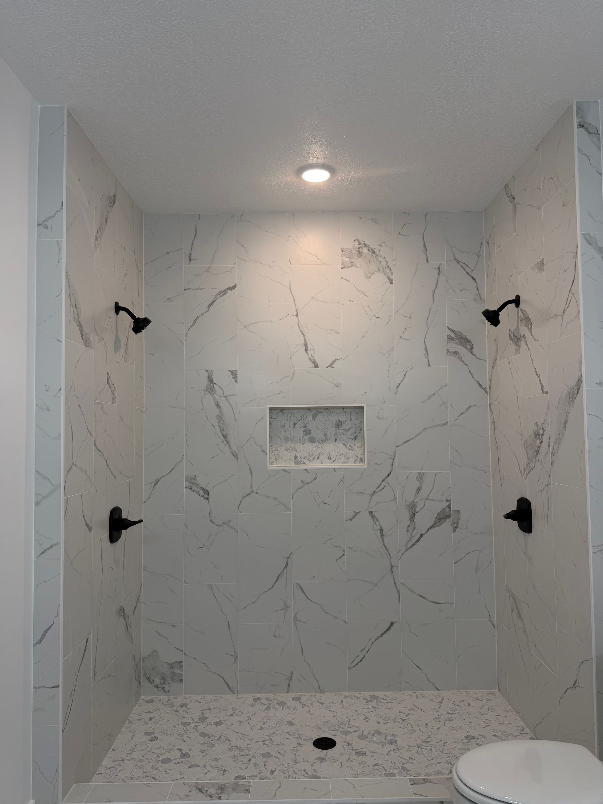 Empty marble-tiled shower stall with recessed shelf and black grab bars.