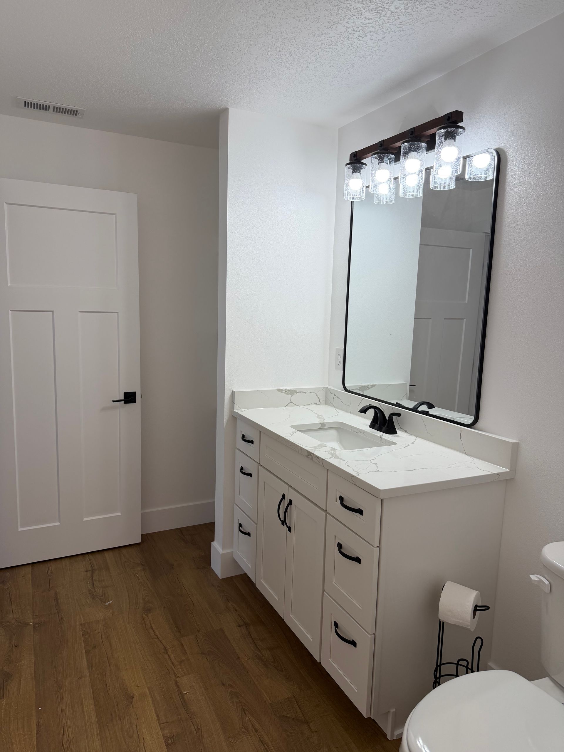 Bright bathroom vanity with mirror, white cabinets, and wood-look flooring.