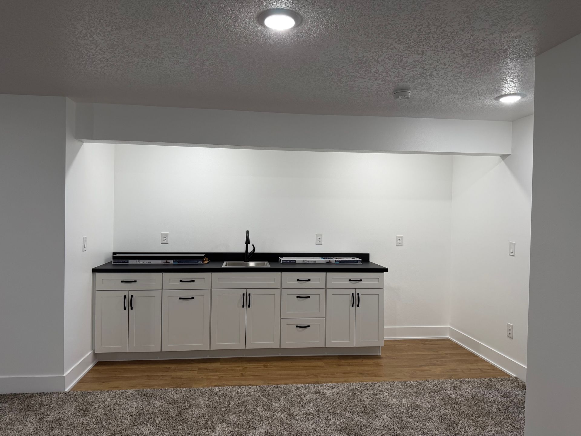 Empty kitchen nook with white cabinets, black countertop, and wood flooring under recessed lights