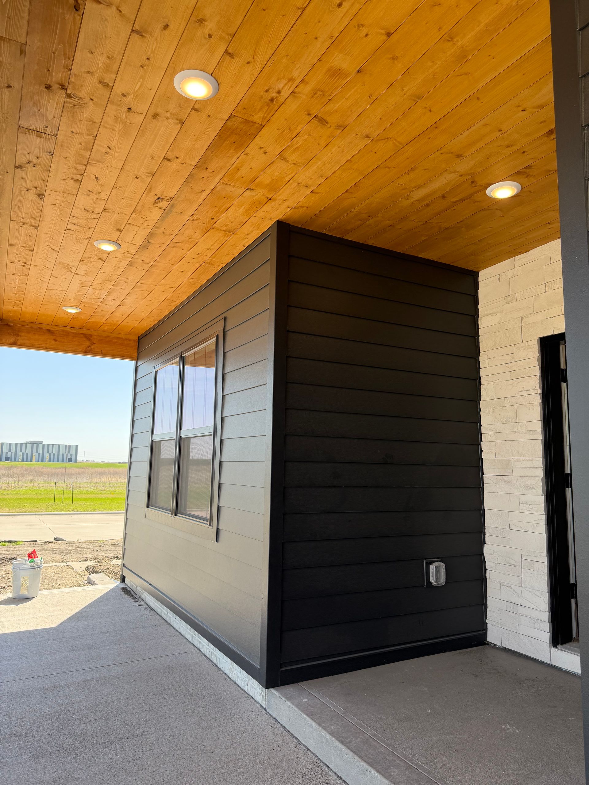 Modern exterior walkway with black siding, wood ceiling, windows, and an open view to the outdoors