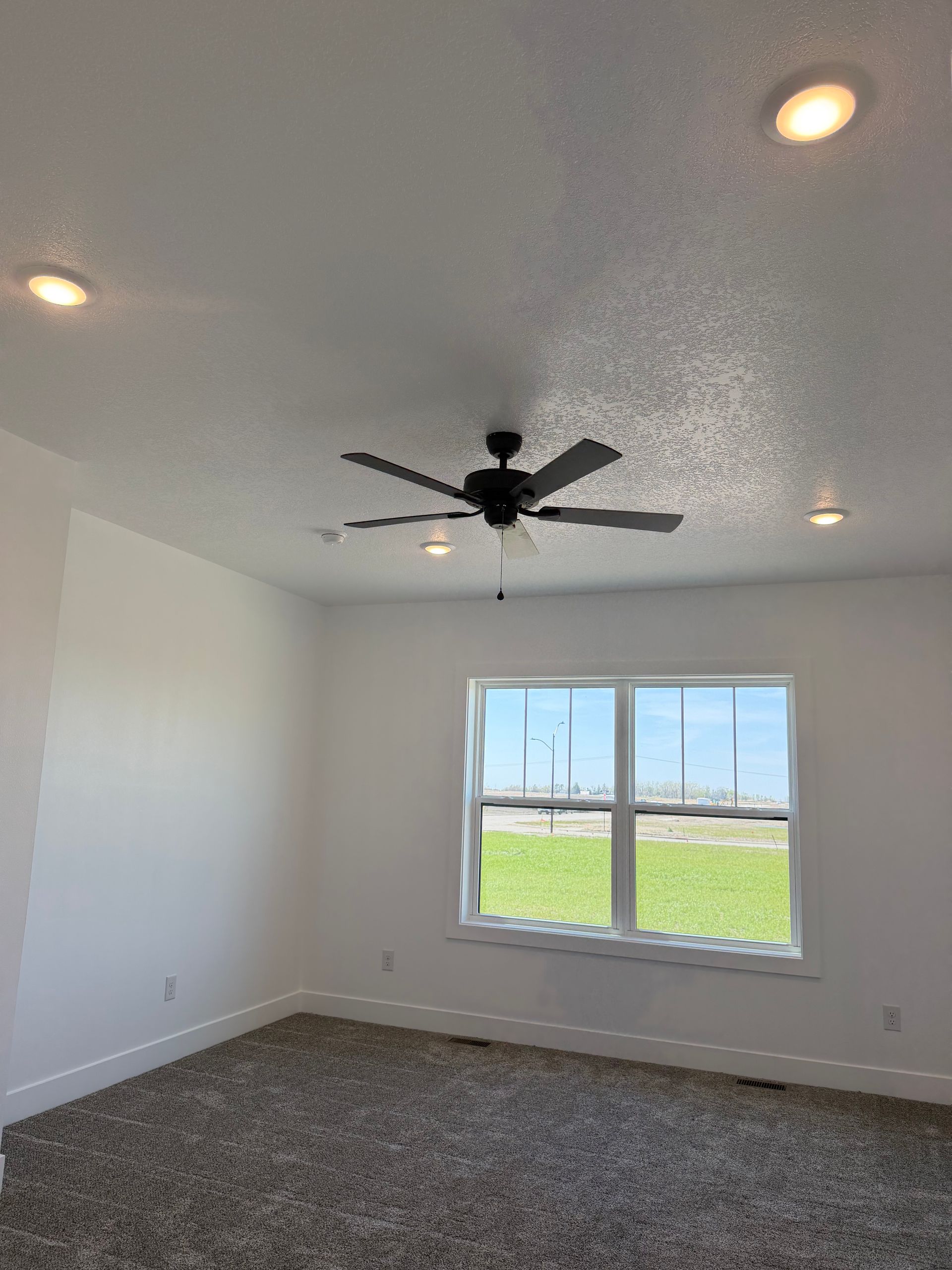 Empty white bedroom with ceiling fan, recessed lights, window, and gray carpet.