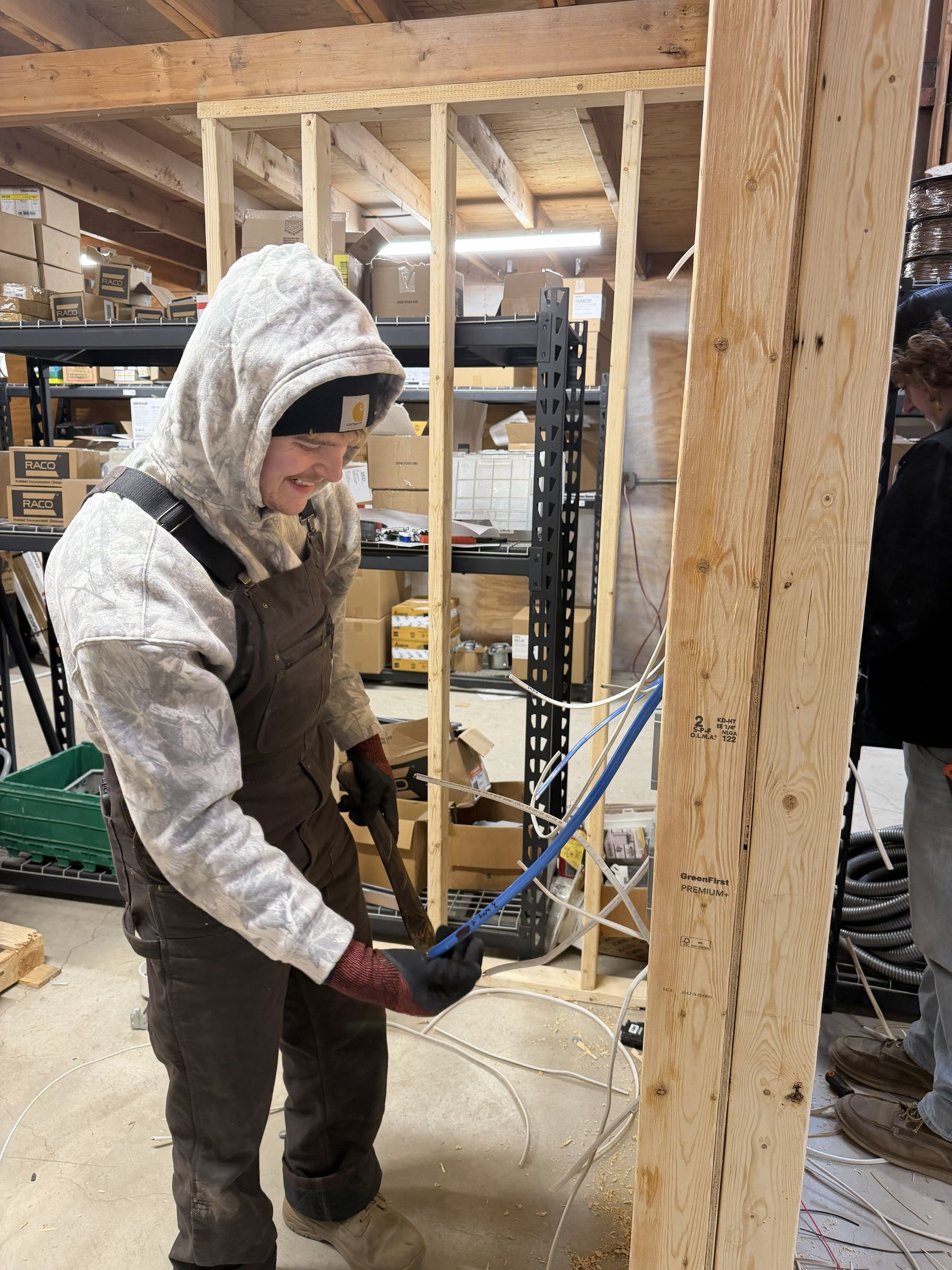 Person in hooded sweatshirt cutting blue pipe in wooden structure. Shelves with items in background.