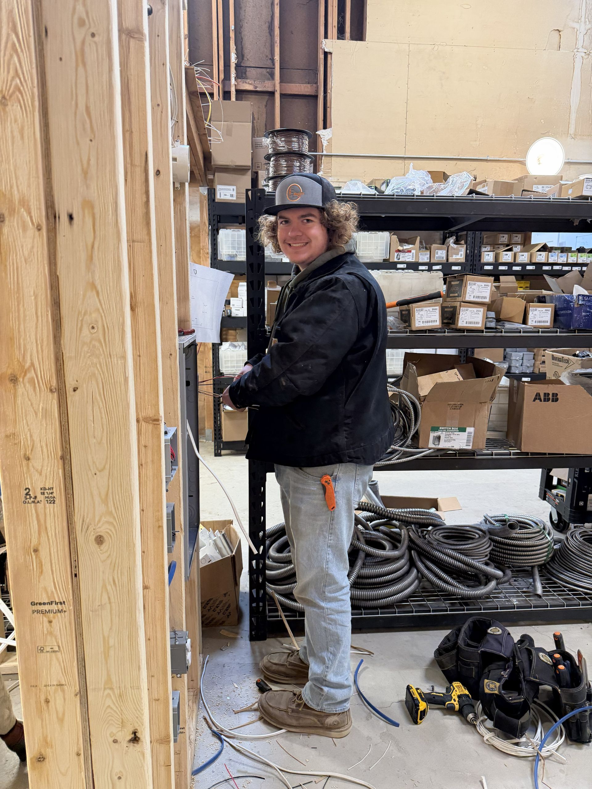 Electrician works on wiring in a construction site. Wearing work clothes, smiling. Wooden studs and shelves with parts in the background.