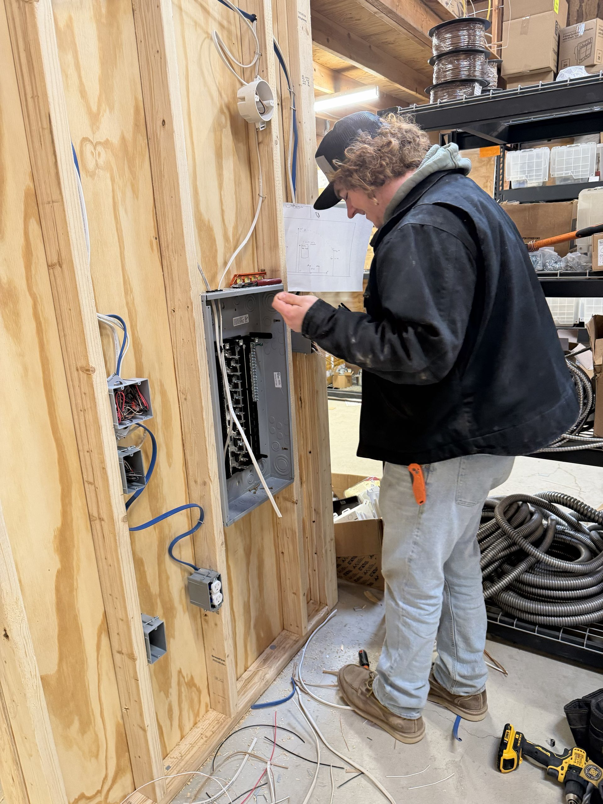 Electrician working on a circuit breaker panel mounted to a wooden wall. Wires and tools are visible.