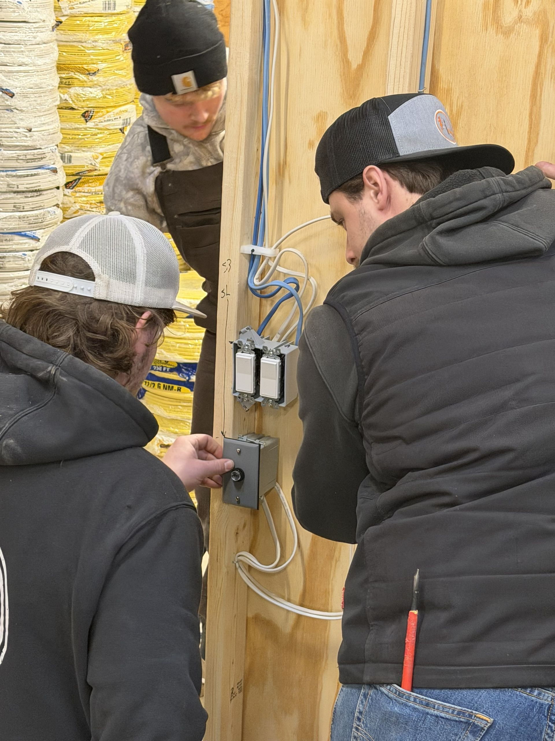 Three people wiring electrical components on a wooden structure. One adjusts a knob, while others look on.