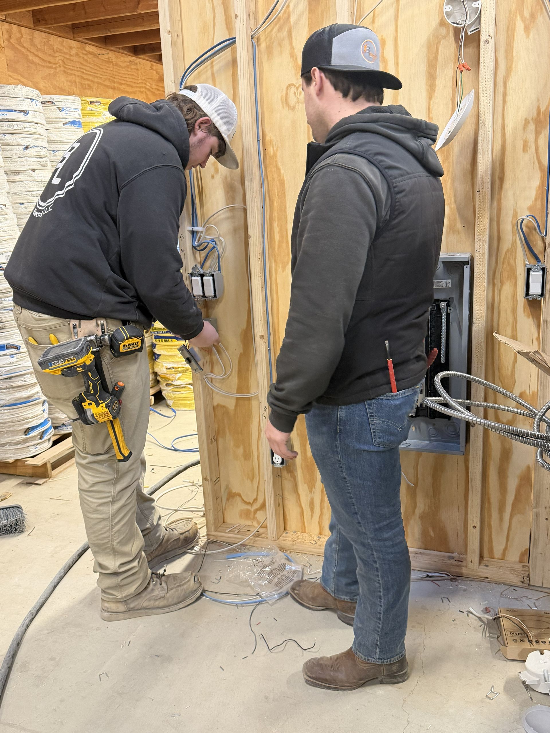Two electricians wiring electrical boxes in a wooden wall. One works, another observes.