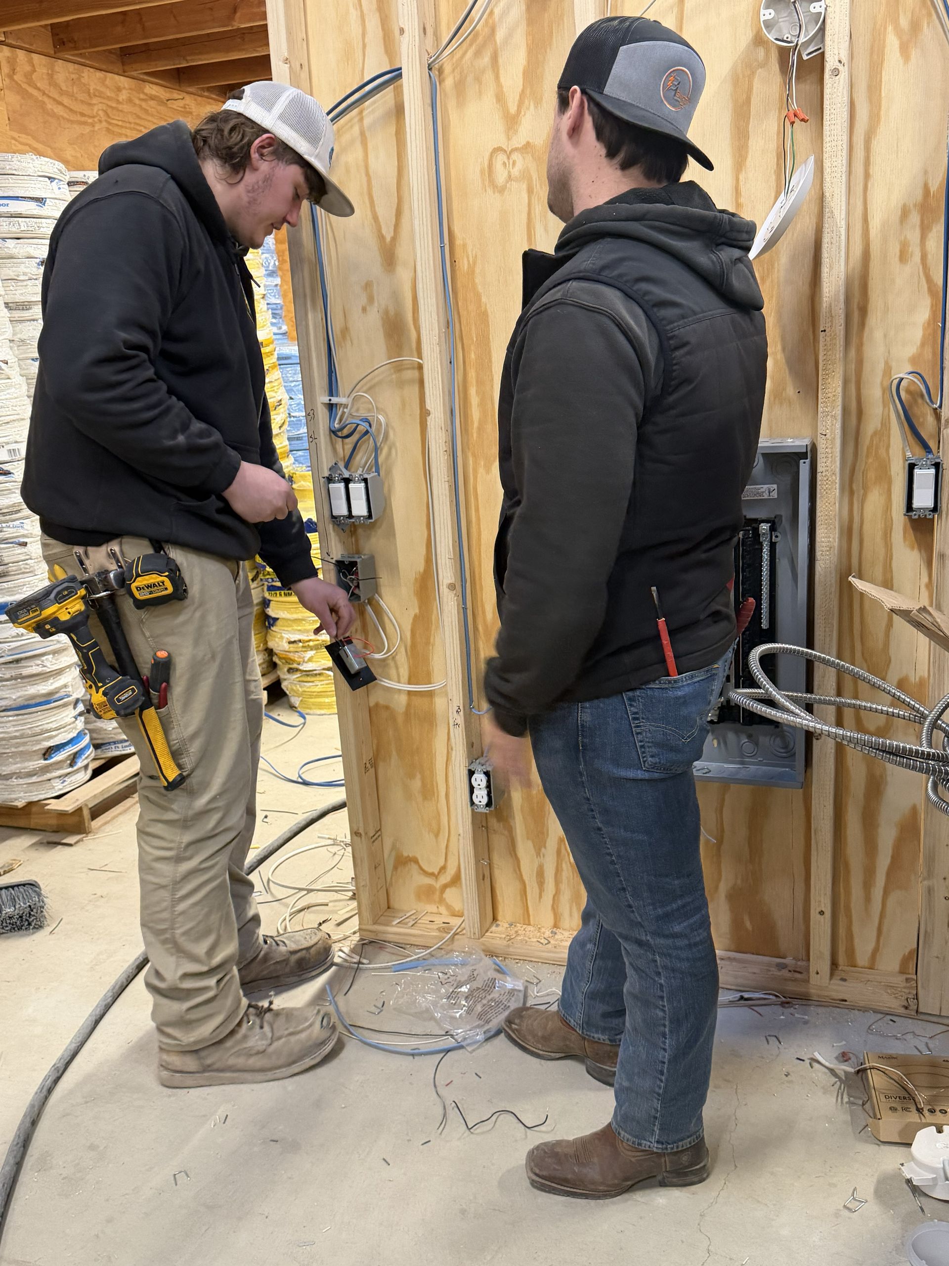 Two electricians working on wiring in a wood-framed structure. One examines wires, other observes.