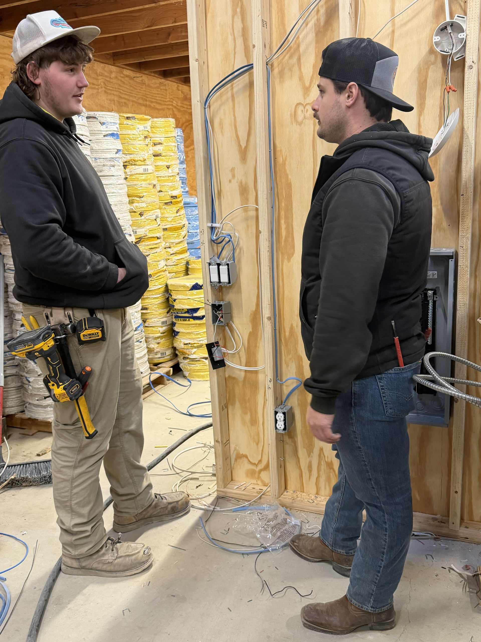 Two electricians stand by wiring in a partially built wall. One in a hat and vest, the other with a tool belt.